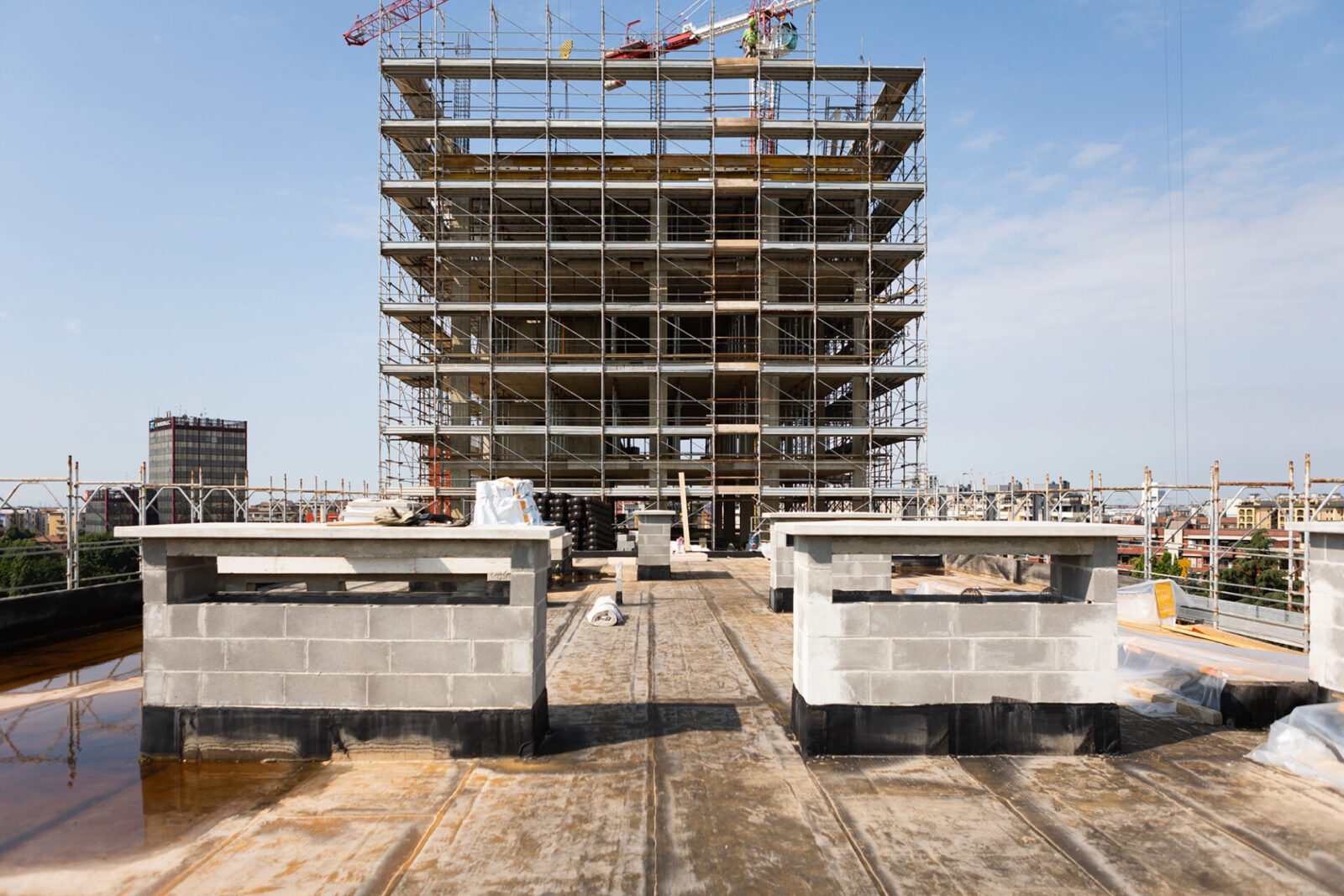 Milano City Village, Milan. Frontal view of a building under construction, surrounded by scaffolding and showing exposed reinforced concrete structure. In the foreground, a rooftop under development with technical components and construction materials. Cranes and the city skyline can be seen in the background.