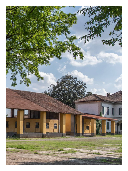 Cascina Ri-Nascita, Milan. Traditional rural buildings with ochre-colored facades and red-tiled roofs surrounded by greenery, under a bright sky with scattered clouds, representing a quiet countryside setting.
