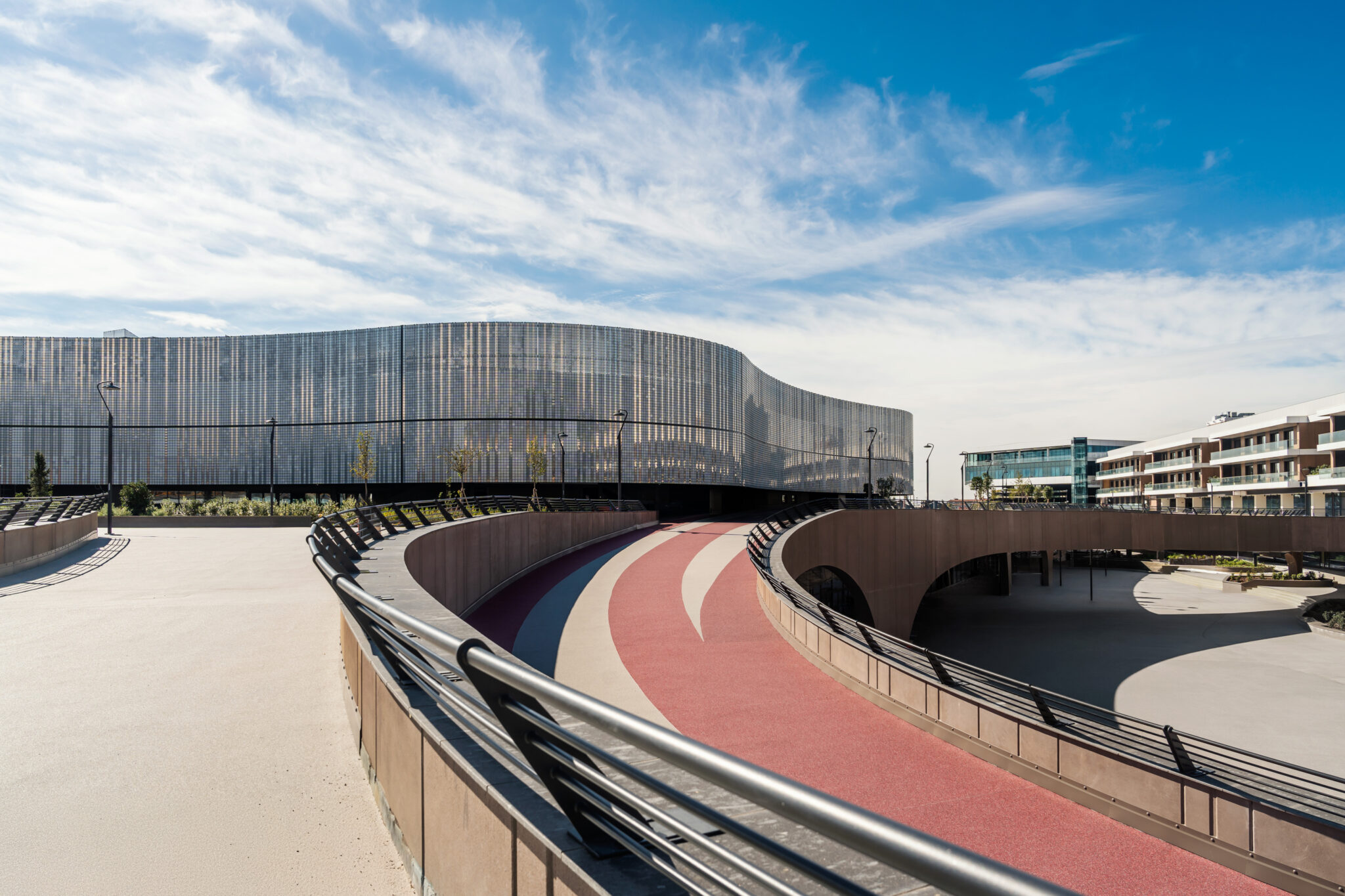 Chorus Life, Bergamo. Curved pedestrian and cycling path with red and white lanes leading to a contemporary building with a wavy, transparent façade under a blue sky with scattered clouds.