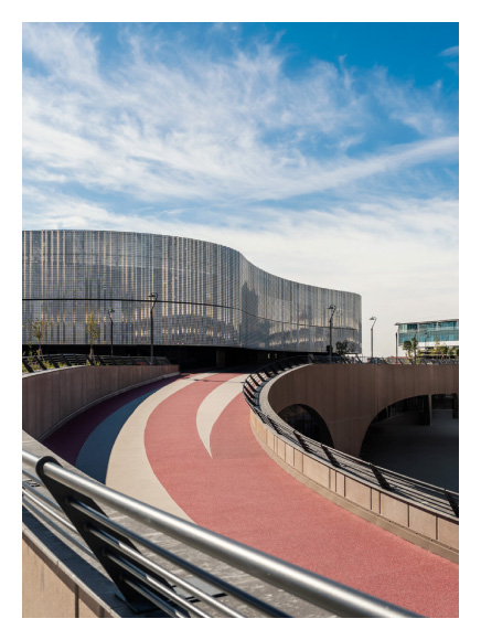 Chorus Life, Bergamo. Curved pedestrian and cycling path with red and white lanes leading to a contemporary building with a wavy, transparent façade under a blue sky with scattered clouds.