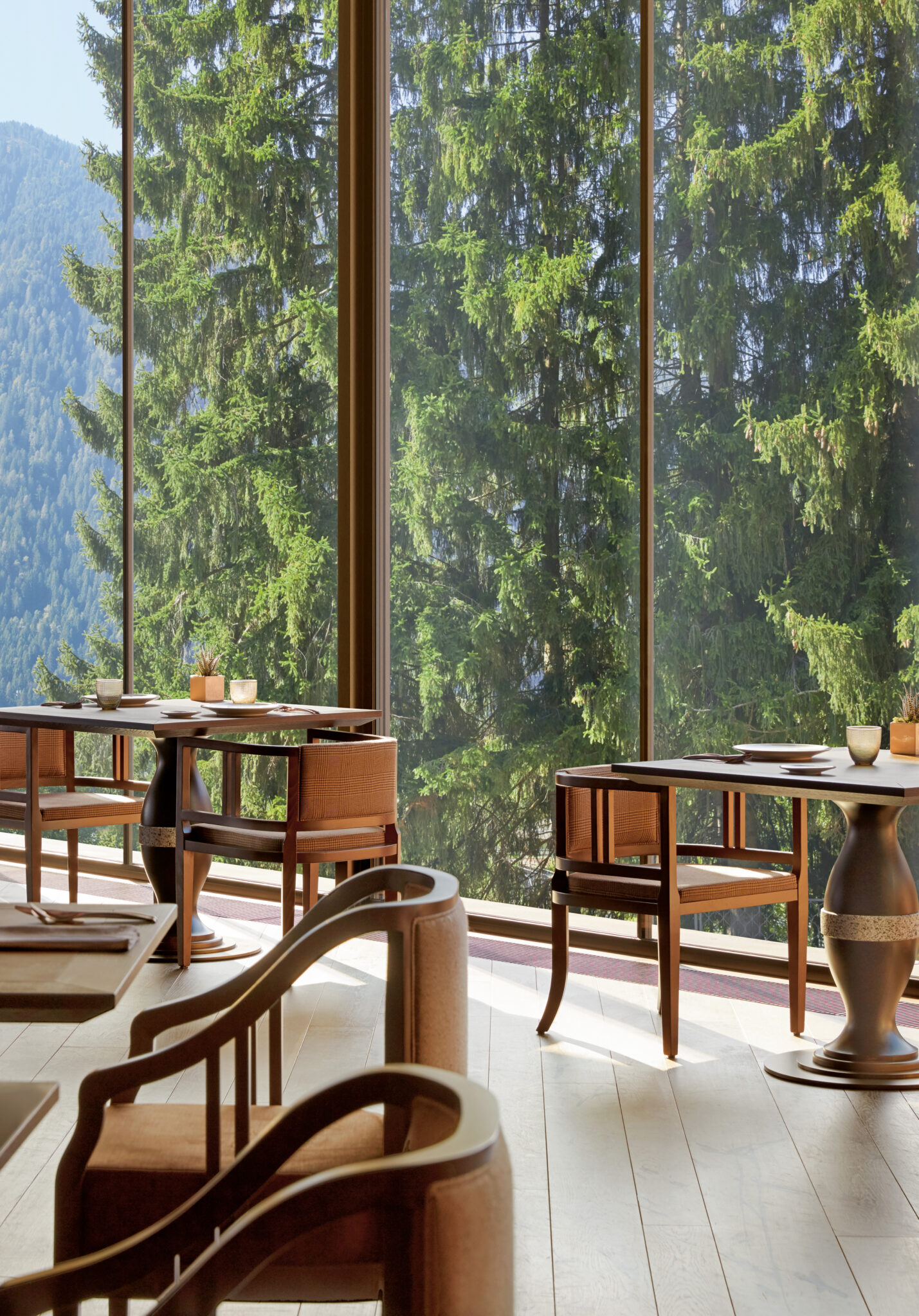 Lefay Dolomites, Pinzolo. Elegant interior of a restaurant with floor-to-ceiling windows overlooking a fir forest. The natural wood furnishings and curved seating design create a warm and refined atmosphere. Natural light filters through the trees, illuminating the light wood floor and the neatly set tables.
