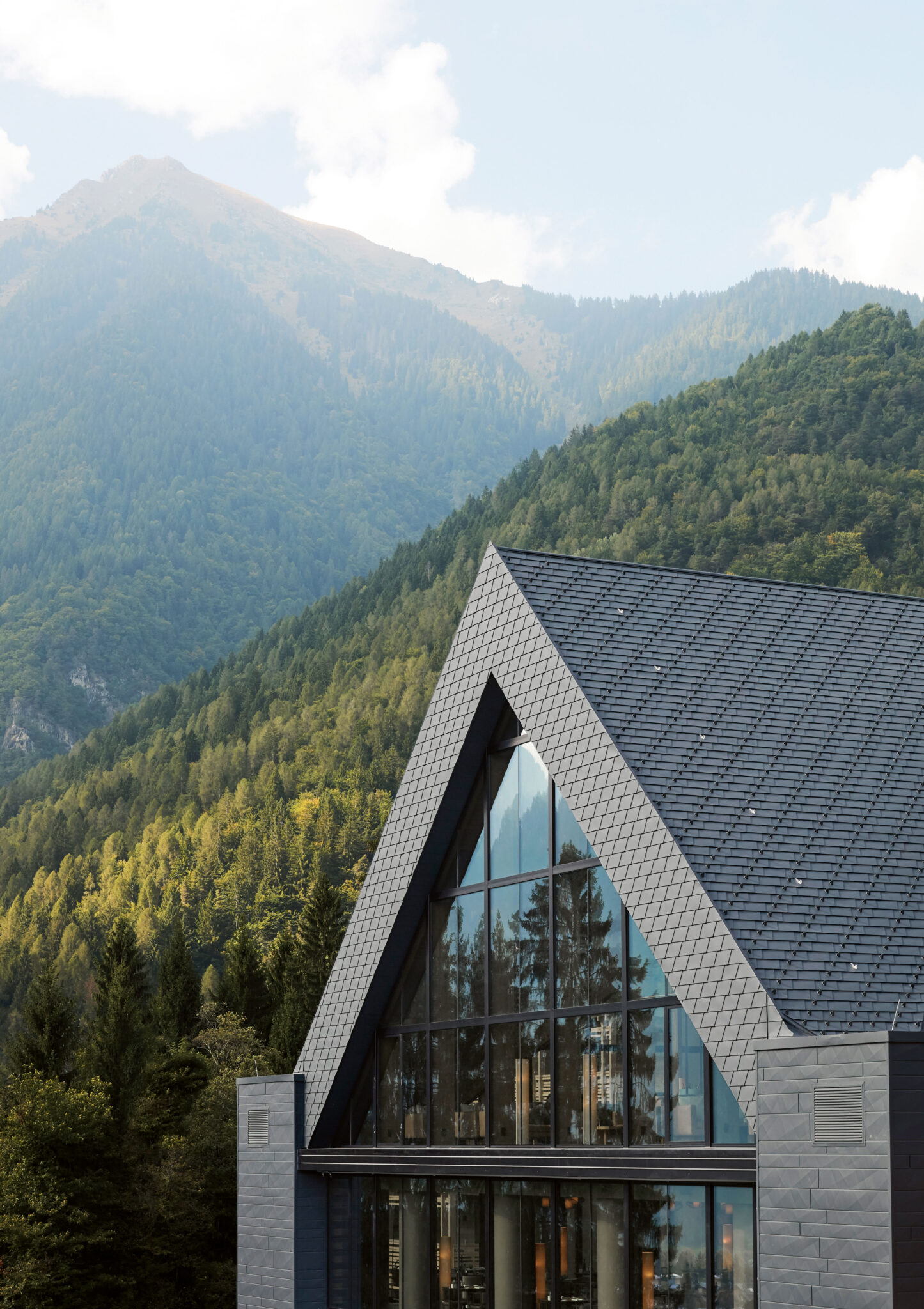 Lefay Dolomites, Pinzolo. Exterior view of the building with a steep gabled roof clad in dark shingles and a full-height glass facade, set against a mountainous landscape with dense forests and scenic peaks.