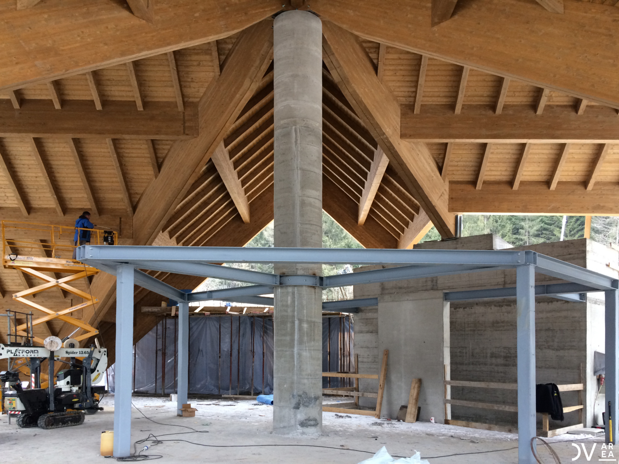 Lefay Dolomites, Pinzolo. Construction phase of a building featuring a pitched roof in laminated wood. At the center, a reinforced concrete column supports a steel frame structure. A worker operates on an elevated platform, while equipment and construction materials are scattered on the ground.