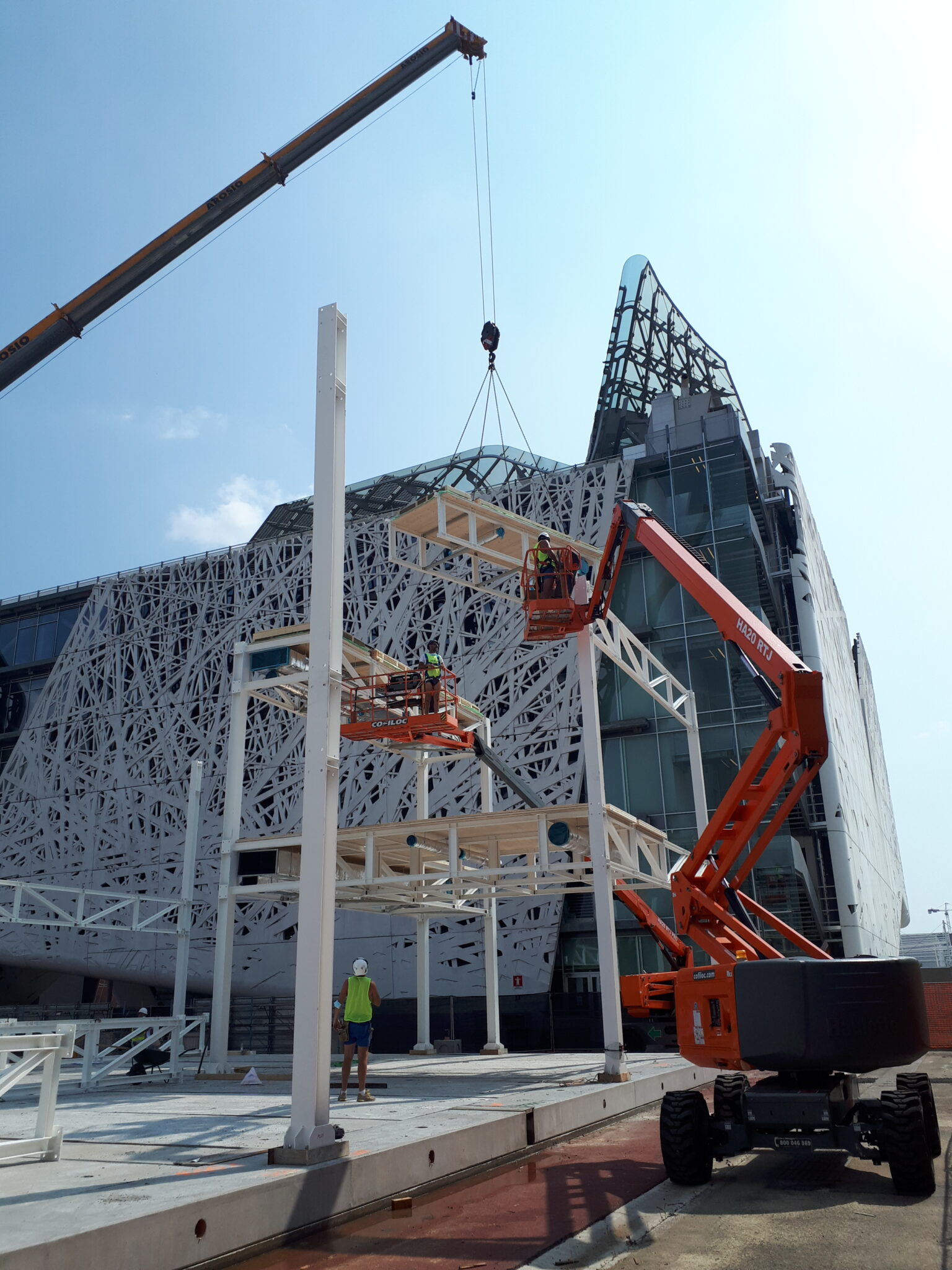 MIND Area, Milan. Assembly of a modular steel structure in front of the Palazzo Italia (EXPO Milan). A prefabricated module is being lifted by a crane while two workers guide it from an aerial platform.