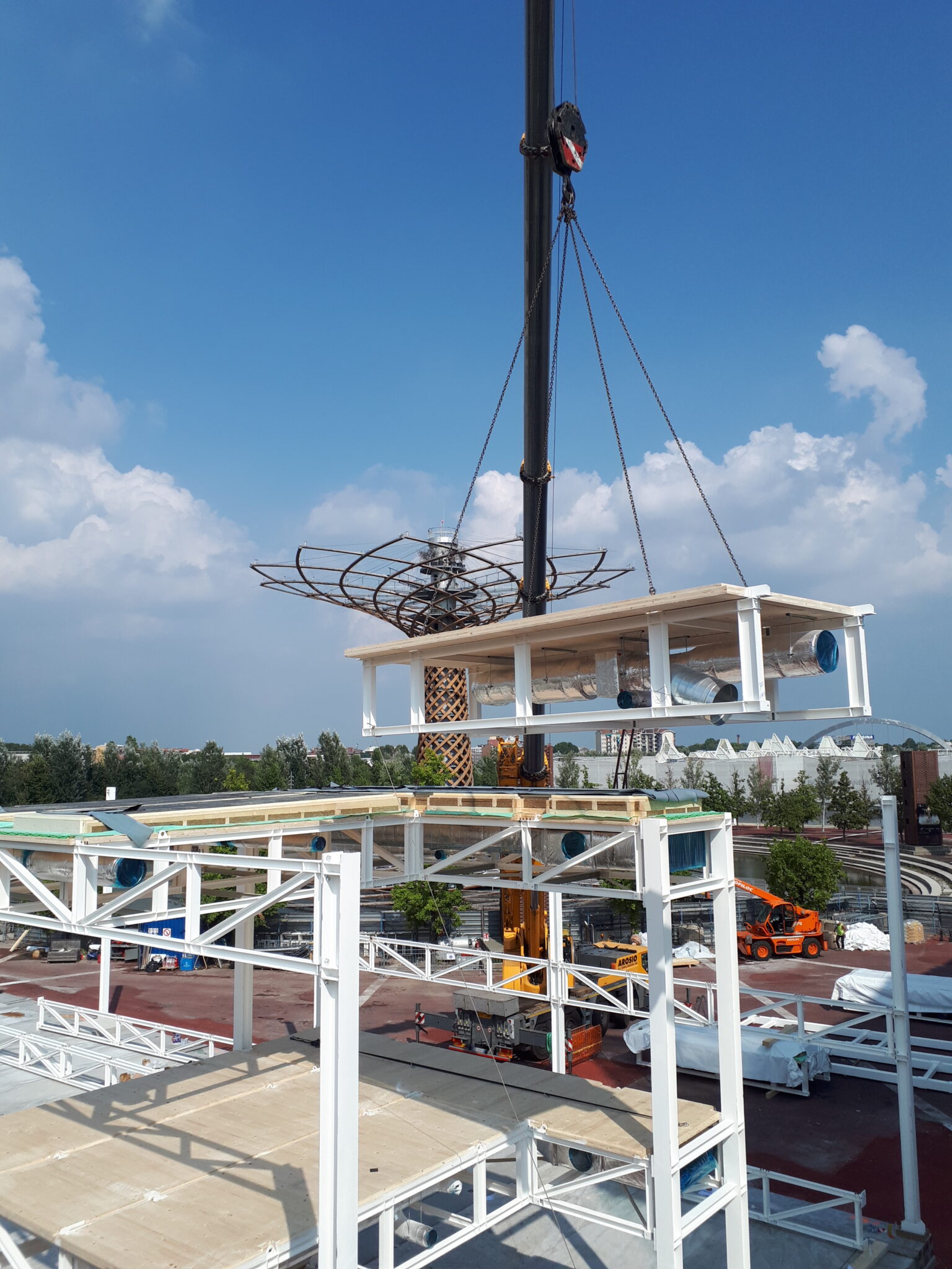 MIND Area, Milan. Lifting of a prefabricated module with integrated systems using a crane at a construction site. The white steel structure is being assembled, with load-bearing elements already in place. In the background, construction machinery, trees, and an urban area are visible.
