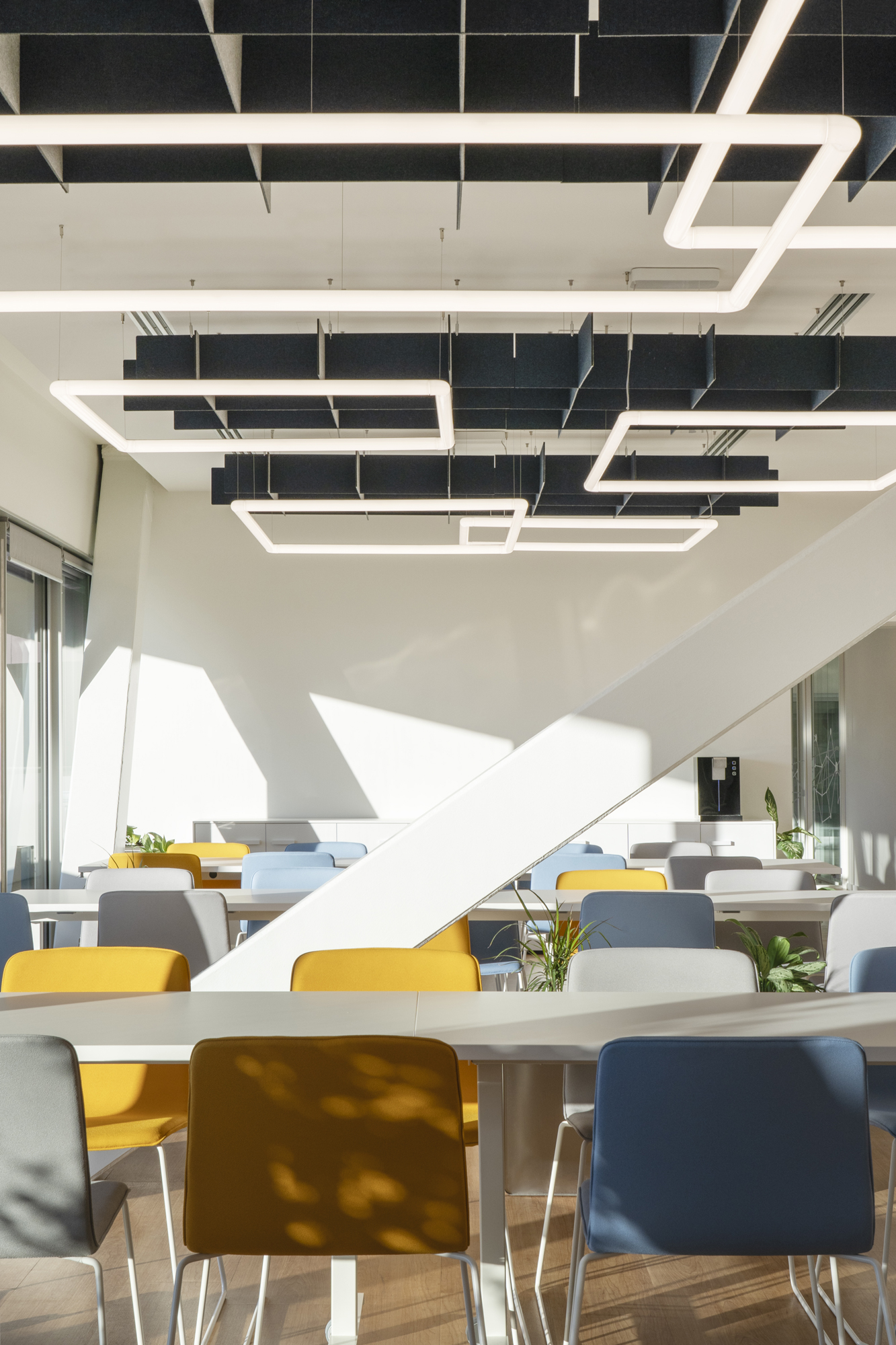 Plenitude + Be Charge Offices, Milan. Bright and modern corporate cafeteria featuring colorful yellow, blue, and grey chairs, white tables, and black acoustic ceiling panels with geometric LED lighting.
