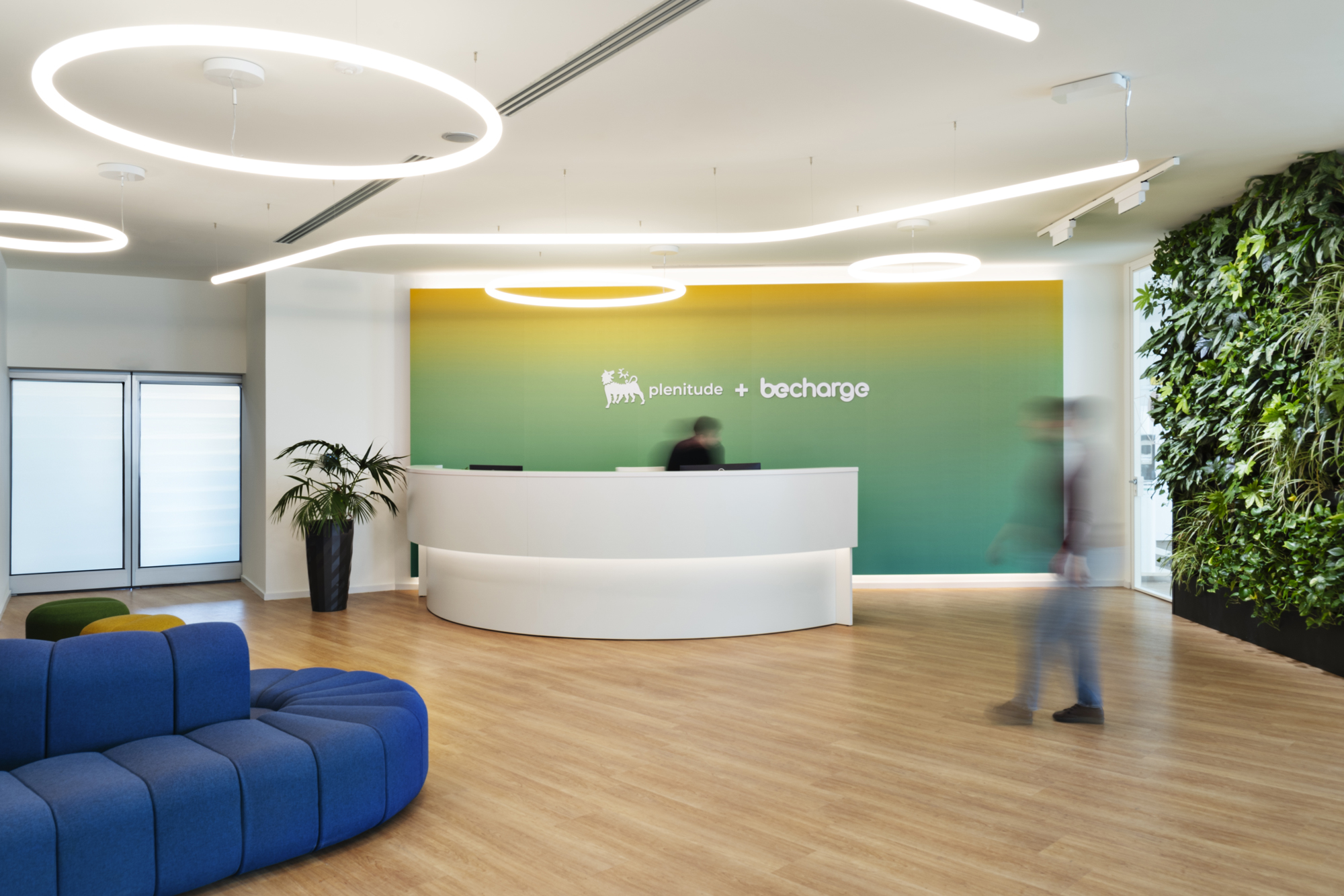 Plenitude + Be Charge Offices, Milan. Reception of the Plenitude + BeCharge headquarters featuring a white curved desk, gradient green-yellow backdrop with company logos, contemporary blue seating, and a vertical green wall with natural plants in a bright, modern space.