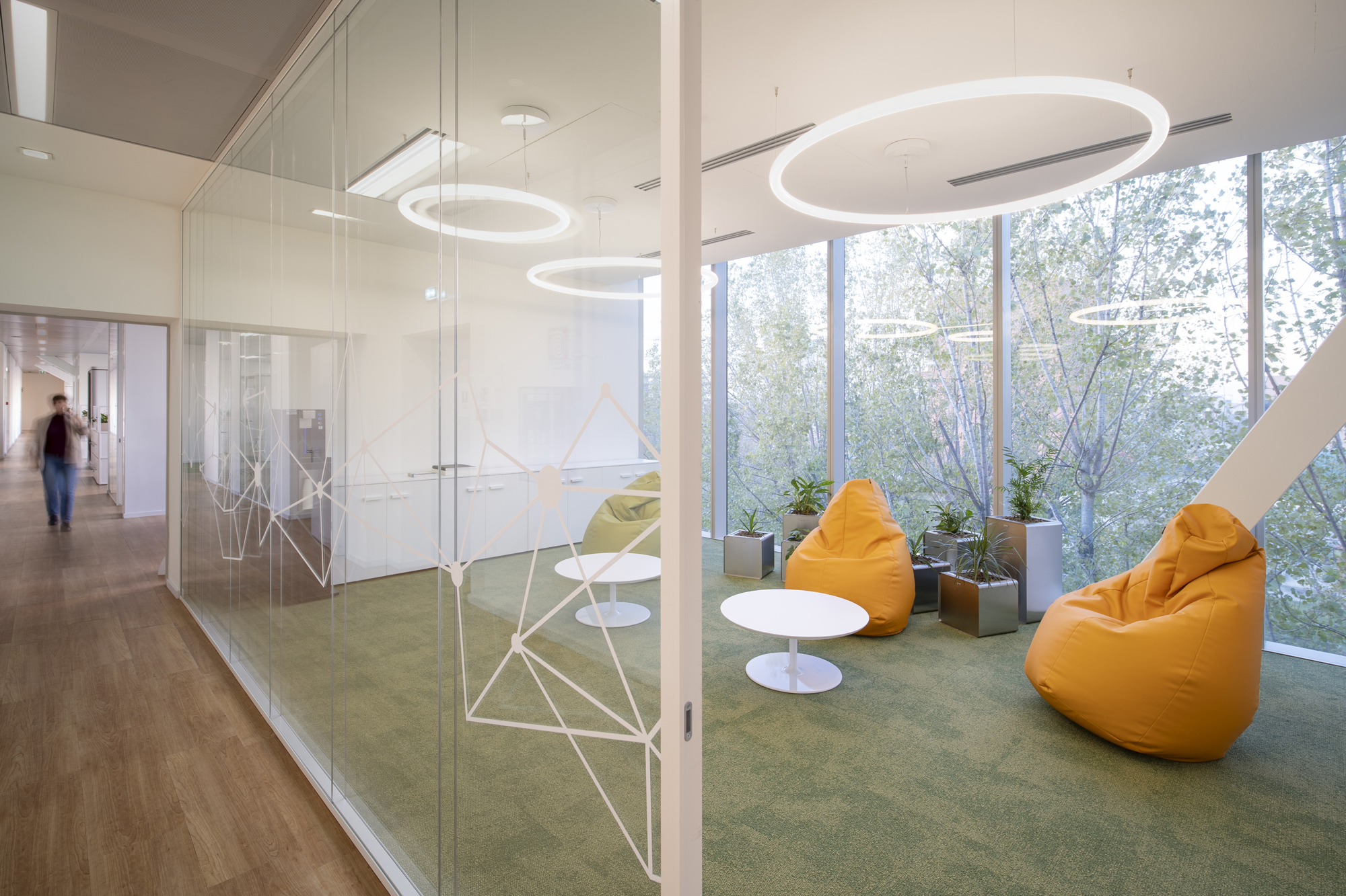 Plenitude + Be Charge Offices, Milan. Relaxation area inside a modern office, with glass walls, large windows facing trees, yellow beanbag chairs, round white tables, and indoor plants.