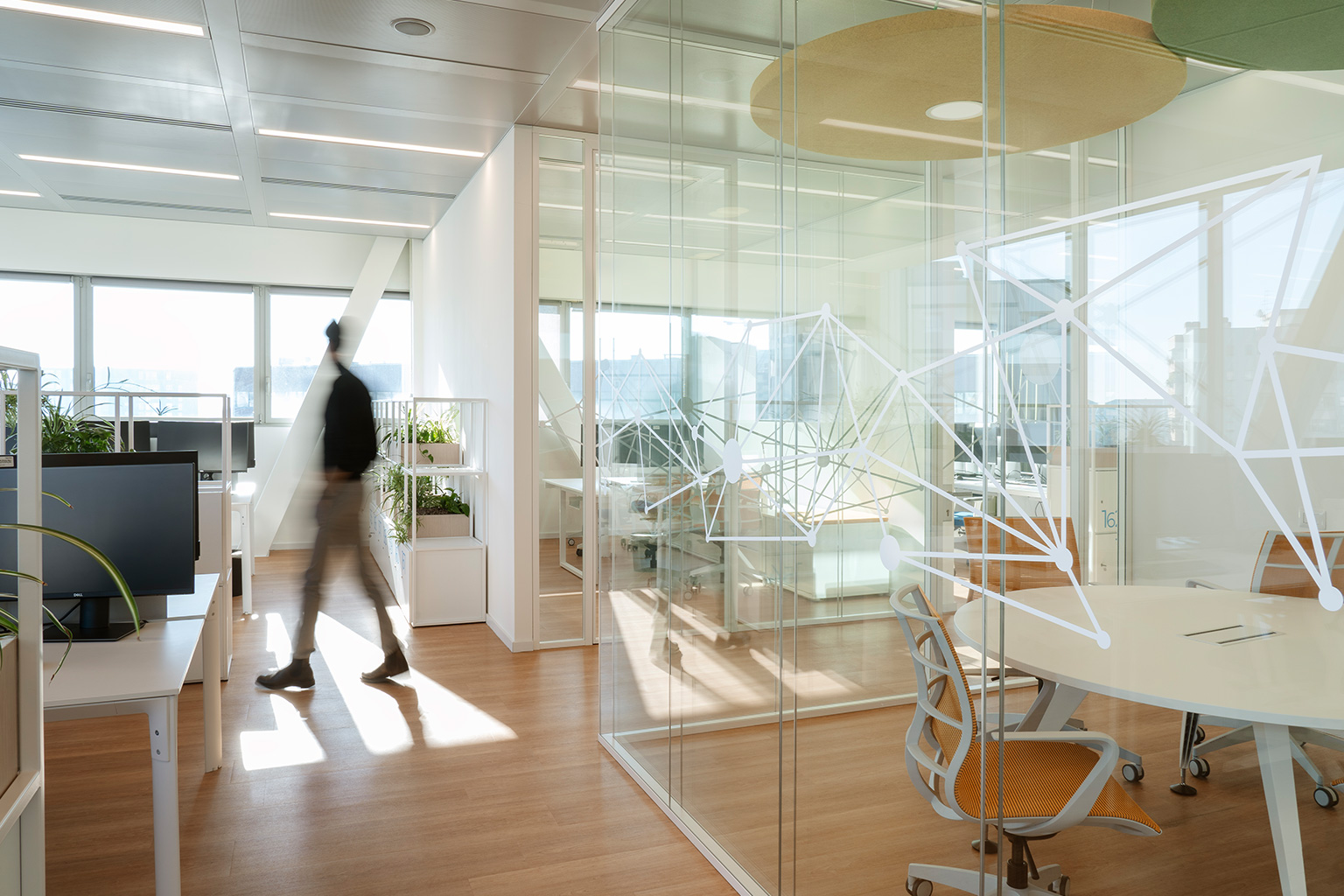 Plenitude + Be Charge Offices, Milan. Modern and bright office space with decorated glass walls, minimalist white furnishings, workstations, and a meeting room with orange ergonomic chairs. A person walks through the corridor.