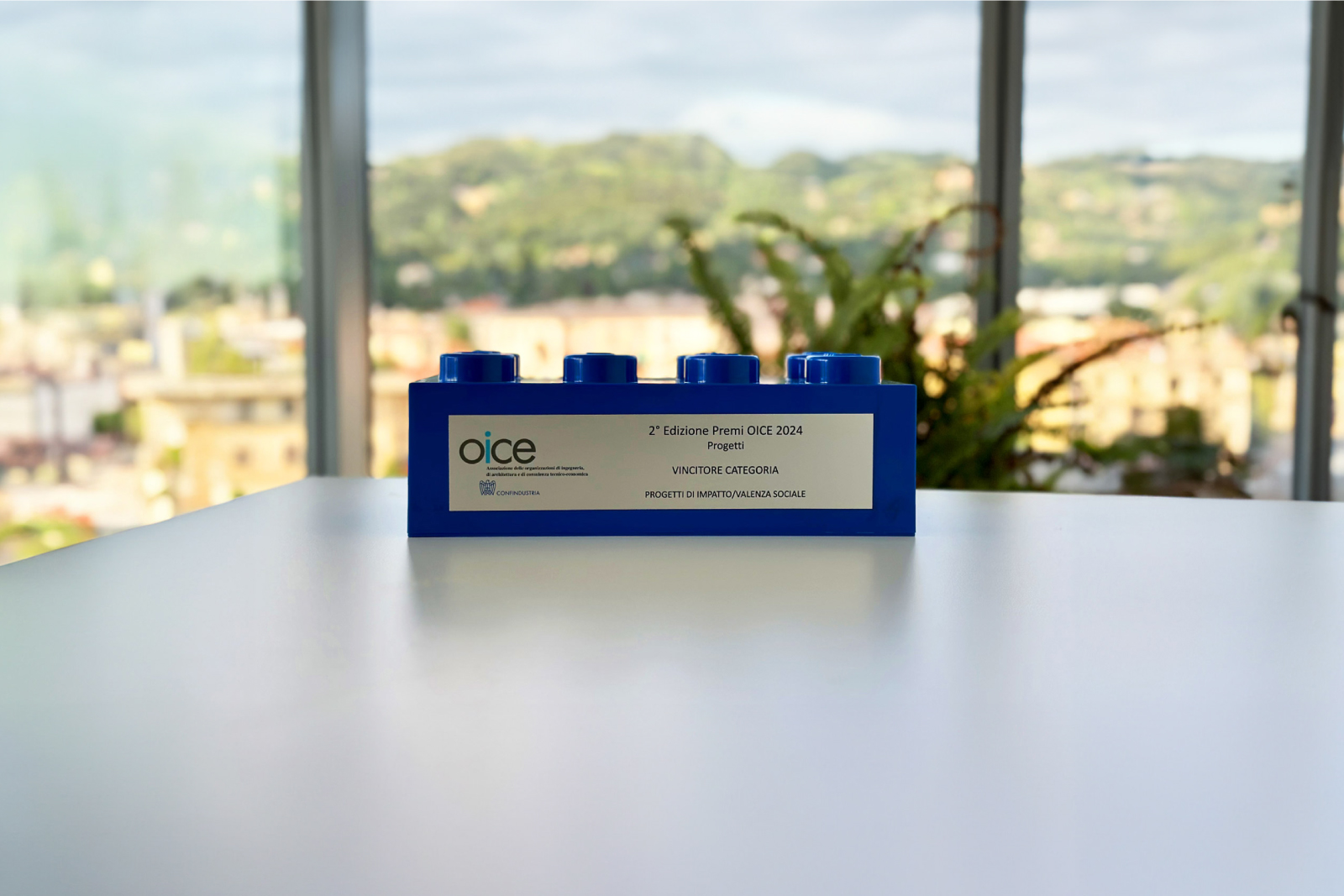 Premio OICE 2024 esposto su un tavolo bianco, con vista panoramica su paesaggio urbano e collinare. Il trofeo a forma di mattoncino blu simile a un blocco Lego celebra la vittoria nella categoria “Progetti di impatto/valenza sociale” durante la 2ª edizione dei Premi OICE, promossi da Confindustria.
