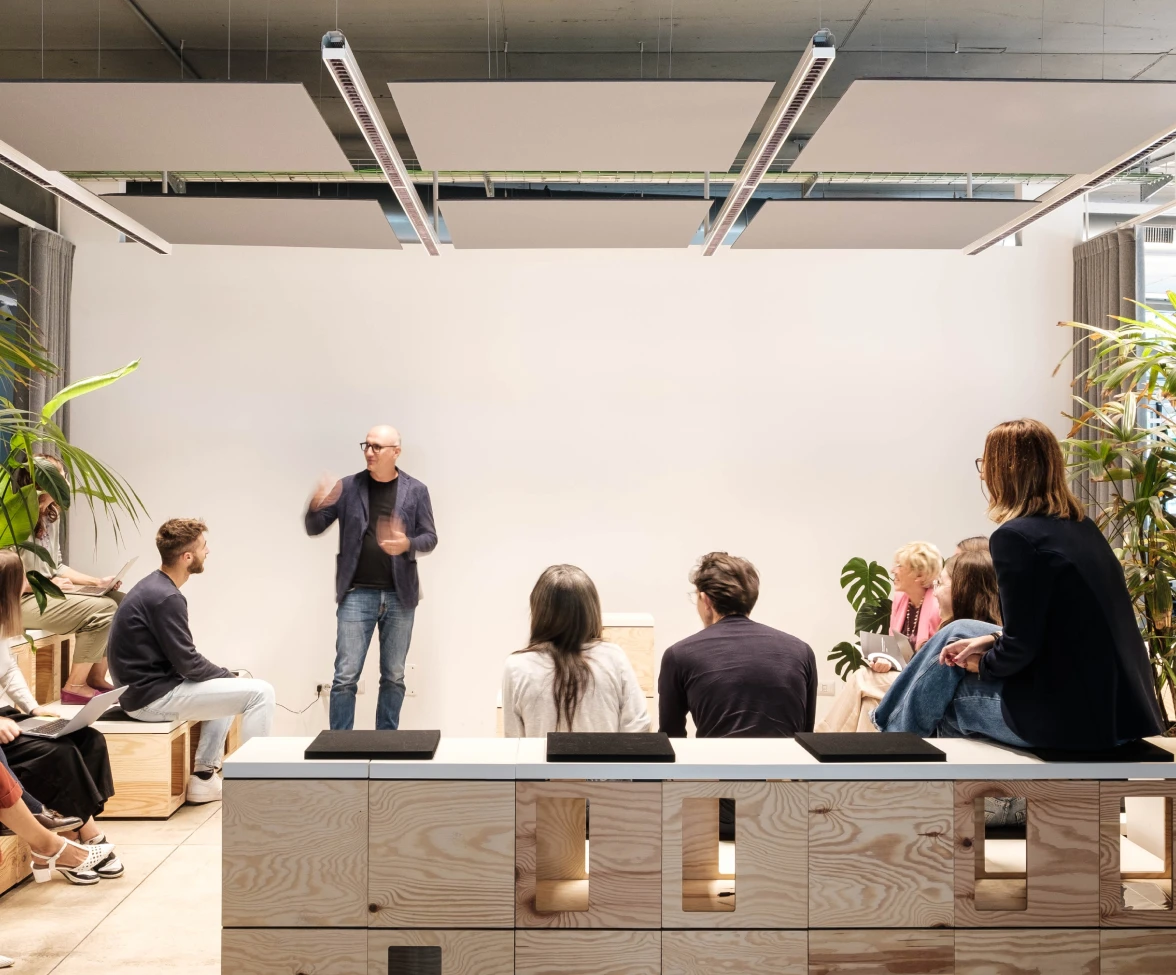 Armando Casella stands while speaking to a group of colleagues seated on modular wooden steps, some with laptops, in an informal room featuring light furnishings, plants, and technical lighting.