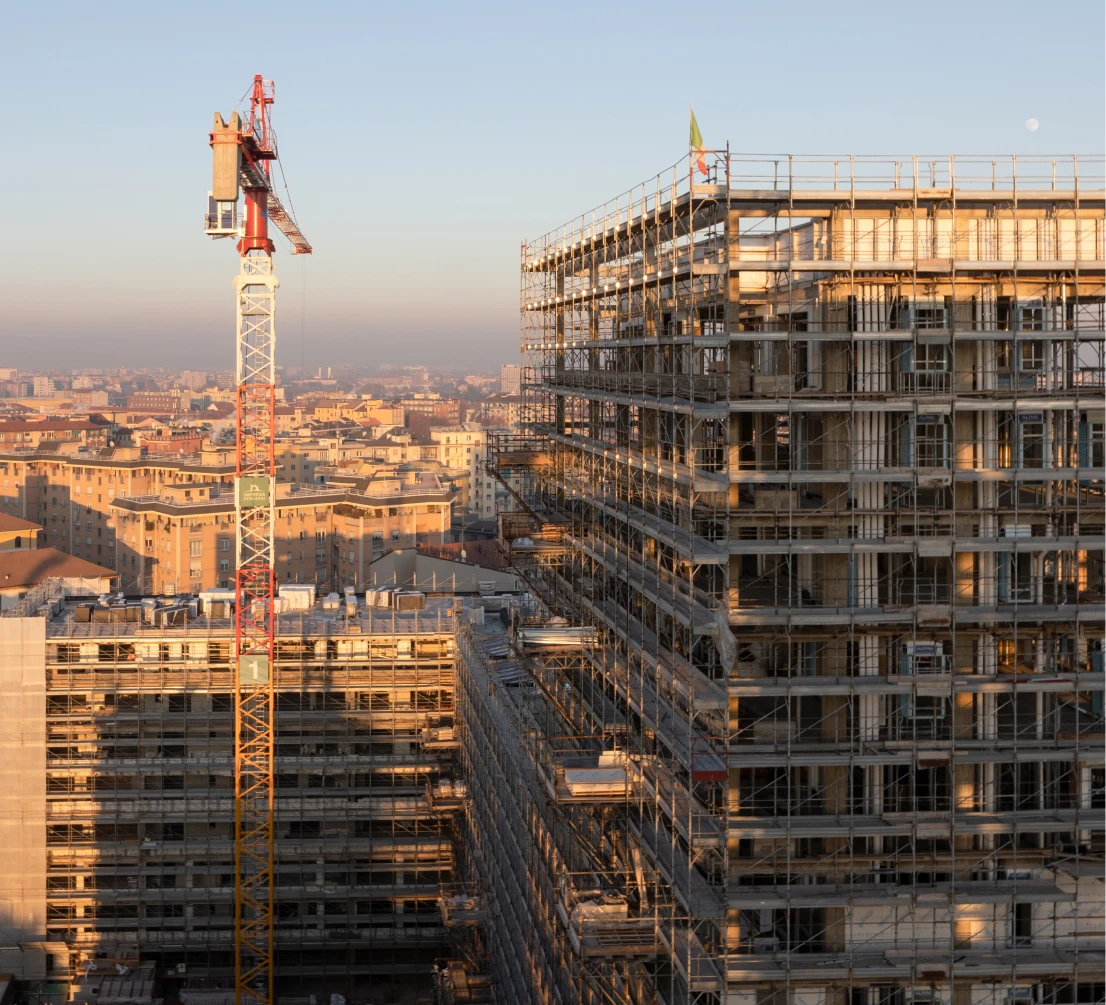 Milano City Village, Milan. Aerial view of the MCV construction site at daybreak, with buildings under construction wrapped in scaffolding, a crane in the foreground, and the urban fabric of Milan in the background.