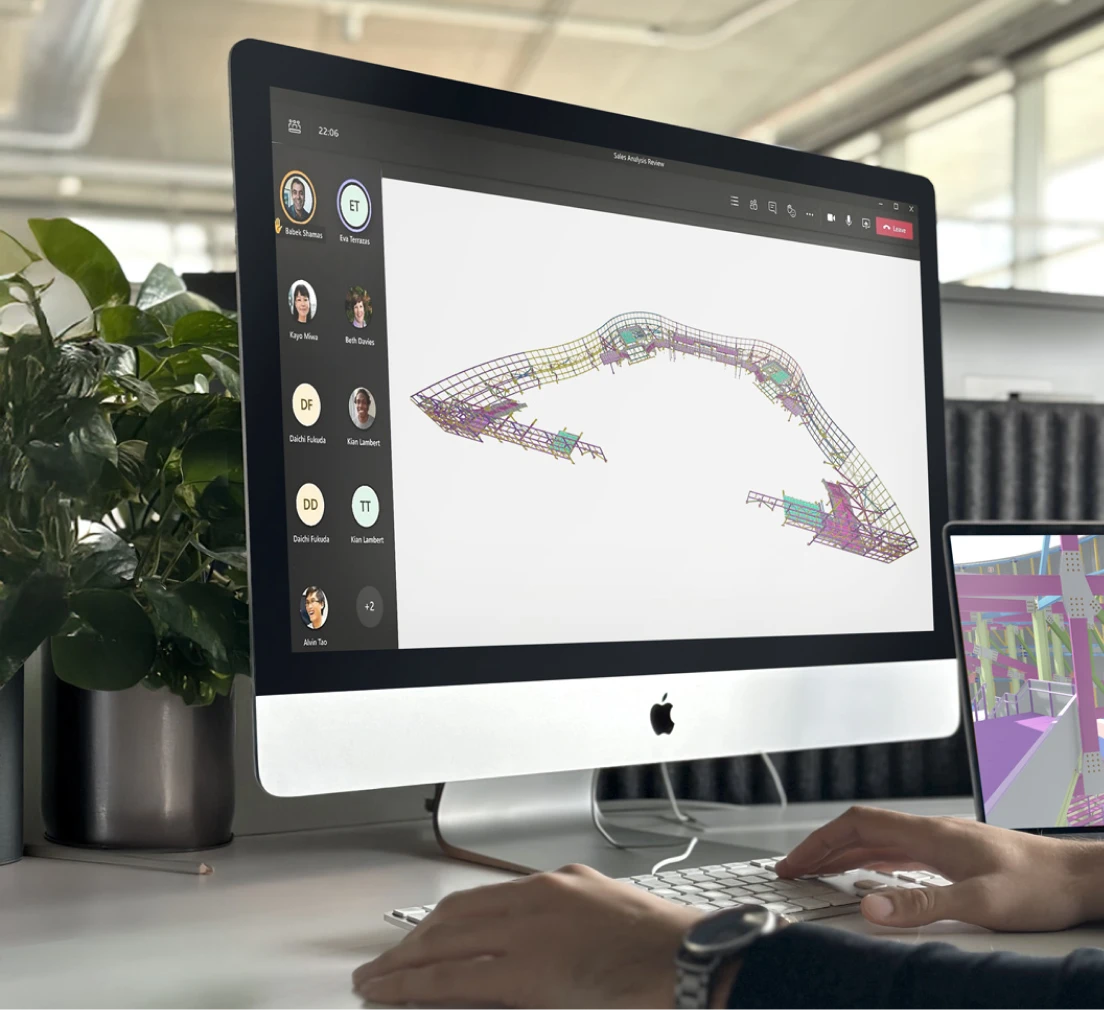 Close-up view of a person working on a computer, with a 3D digital model displayed on the screen during a group video call; a second device on the desk shows a technical visualization, all set in a bright office environment with ornamental plants.