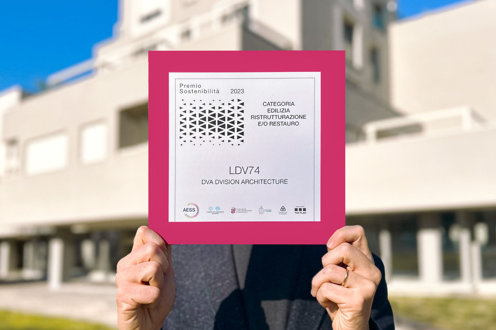 Una persona mostra il Premio Sostenibilità 2023 ricevuto da DVA DVision Architecture per il progetto “LDV74”, vincitore nella categoria “Edilizia, Ristrutturazione e/o Restauro”. Il certificato è incorniciato da un bordo rosa acceso, con sullo sfondo un edificio moderno sfocato.