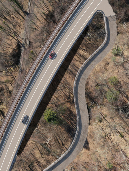 State Road, Val Vigezzo. Aerial view of a road segment immersed in a mountainous forest landscape, with two cars traveling on an elevated viaduct and a new road below following the natural contours of the terrain, near a railway line visible in the lower left corner.