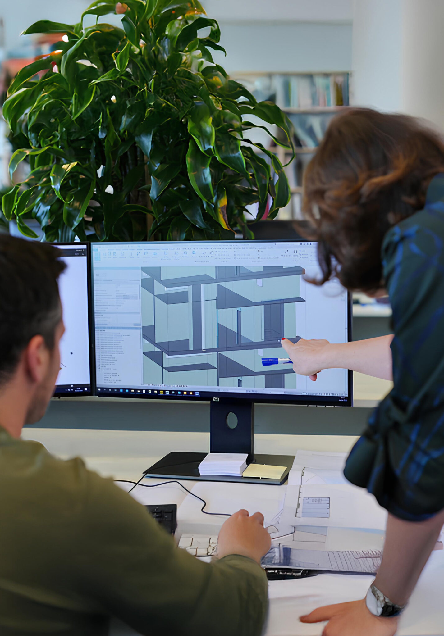 Professionals working on a 3D architectural model using BIM software, displayed on dual monitors in a technical office setting.