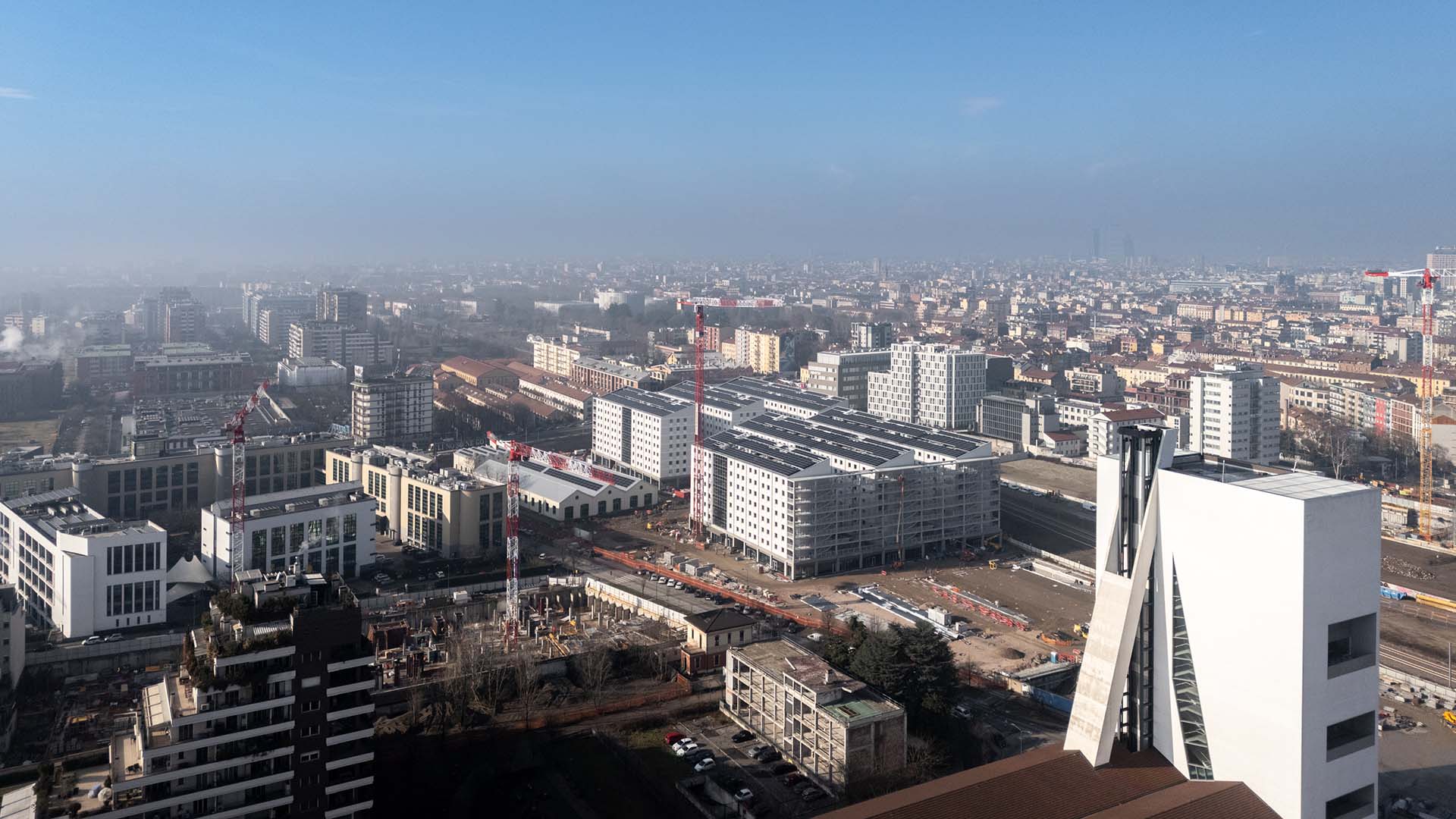 Olympic Village, Milan. Aerial view of the construction site in the Olympic Village, with new residential buildings, skyscrapers, and cranes visible against the background of Milan, in an urban development area.