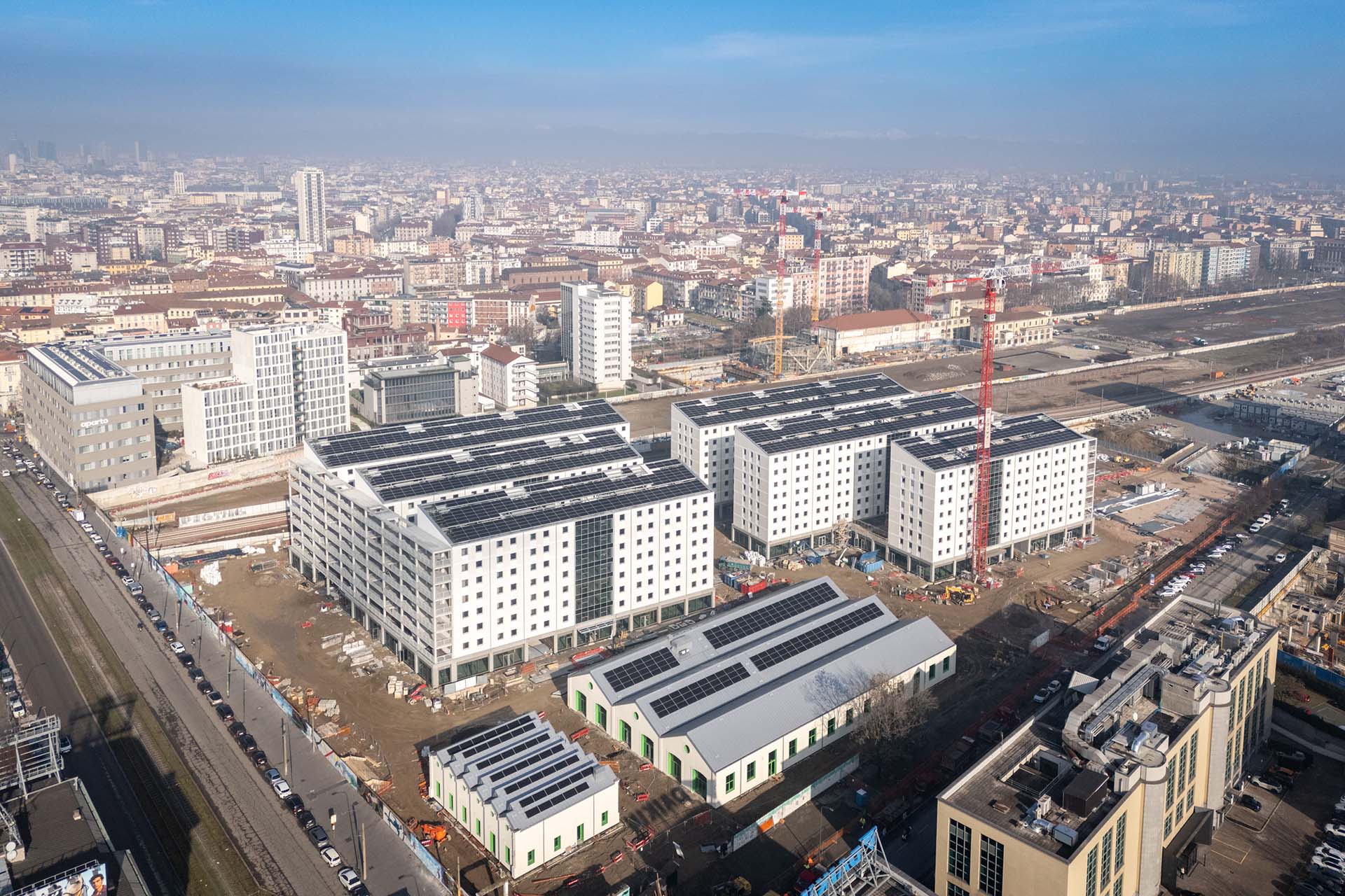 Olympic Village, Milan. Aerial view of the residential complex under construction with solar panels on the roofs, consisting of buildings with modern white facades, located within a developing area, with the construction site visible and the city in the background.