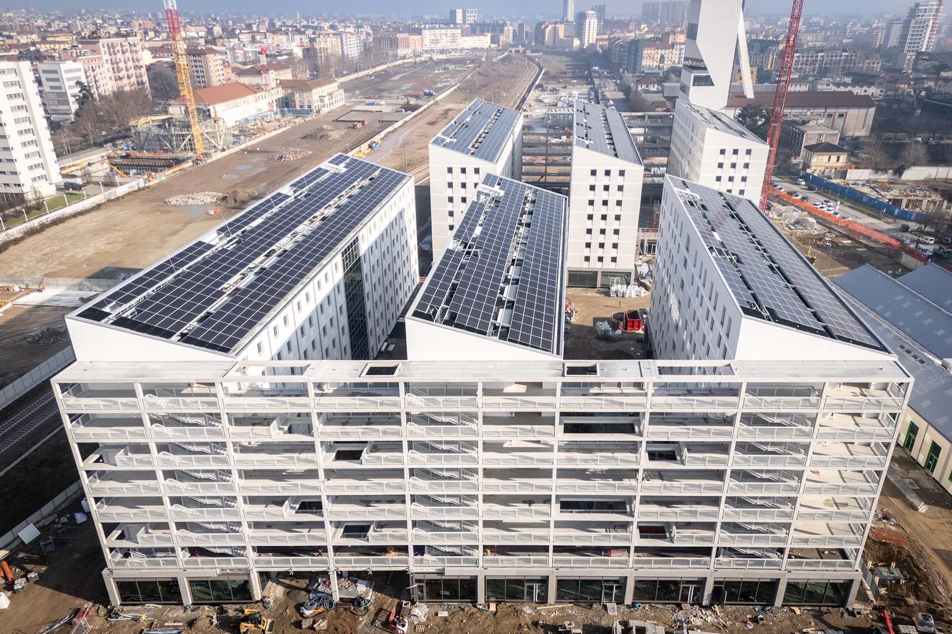 Olympic Village, Milan. Aerial view of the residential complex under construction with solar panels on the roofs, consisting of modern white-facade buildings, located within a developing area, with the construction site visible and the city in the background.