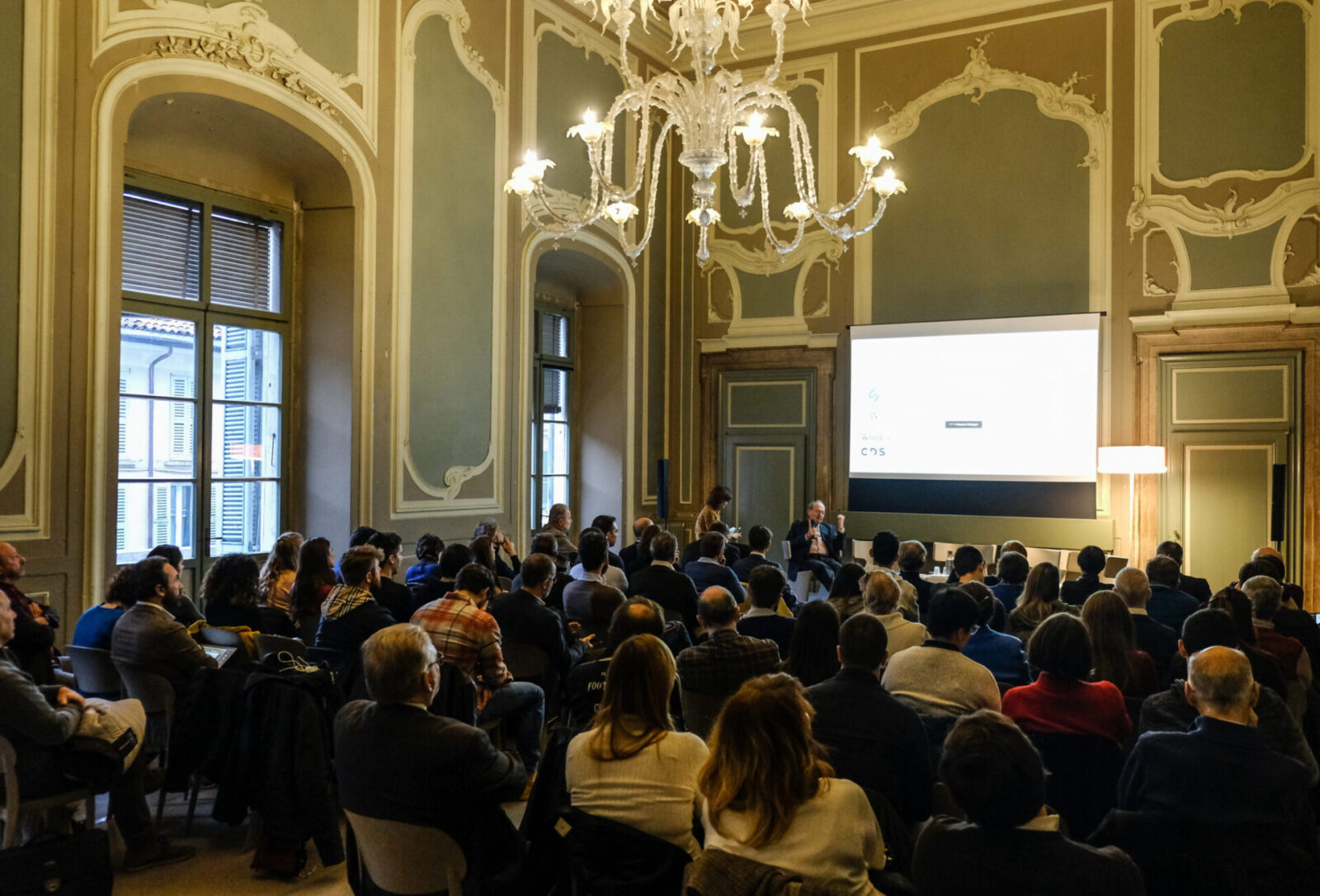 Conference in progress inside a historic room adorned with stuccoes and a glass chandelier. A large, attentive audience is facing a seated speaker in front of a projection screen. The event takes place in an elegant, culturally rich setting—ideal for high-profile gatherings.