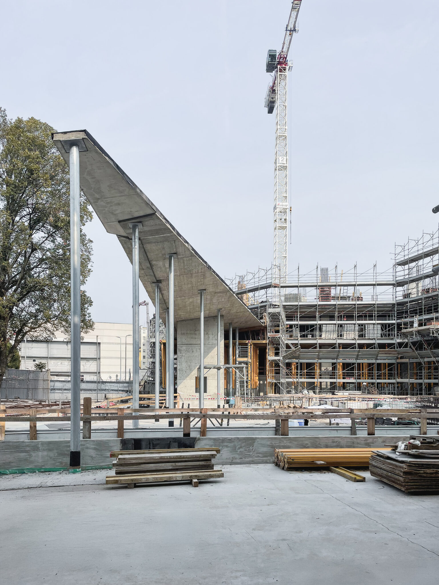 BoscoNavigli, Milan. Construction site with a reinforced concrete structure under development, featuring a sloped roof supported by metal columns, with scaffolding and a crane in the background.