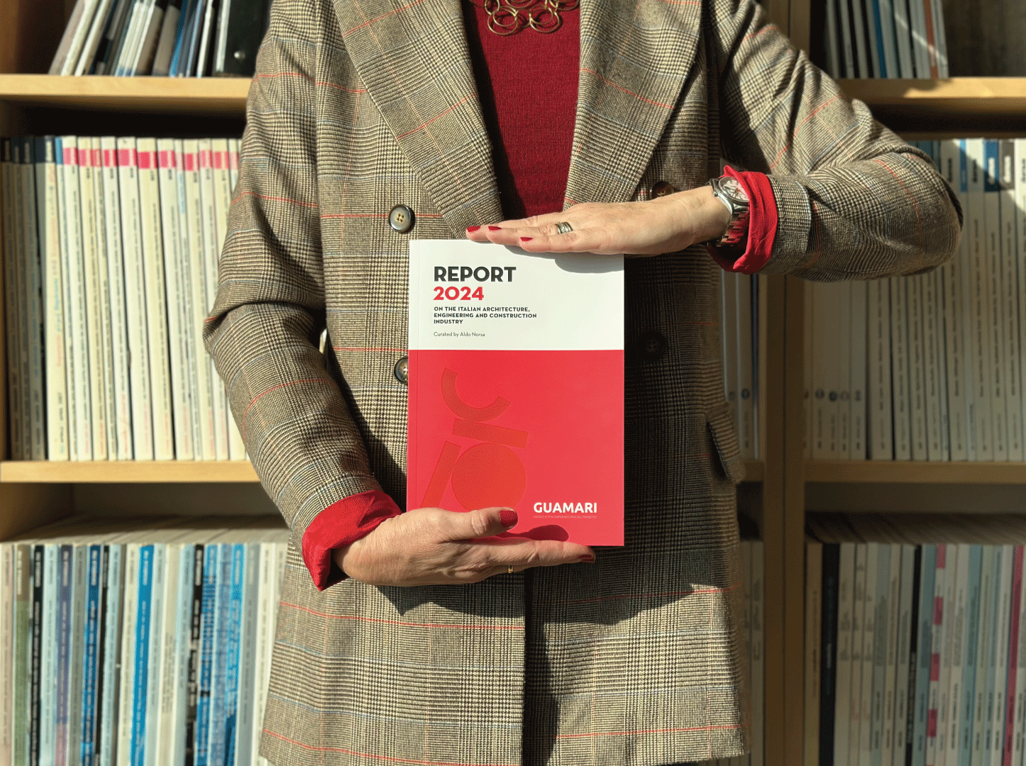 Persona in abiti formali tiene in primo piano una copia del “Report 2024” sul settore italiano dell’architettura, ingegneria e costruzioni, pubblicato da Guamari. Lo sfondo mostra scaffali pieni di riviste tecniche, in un contesto ordinato e professionale.