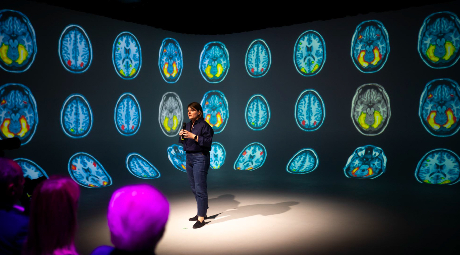 Circolo delle Imprese, Milan. Marta Olivieri on stage presenting in front of an immersive projection of multiple brain scan images, in a dimly lit auditorium with an audience watching attentively.