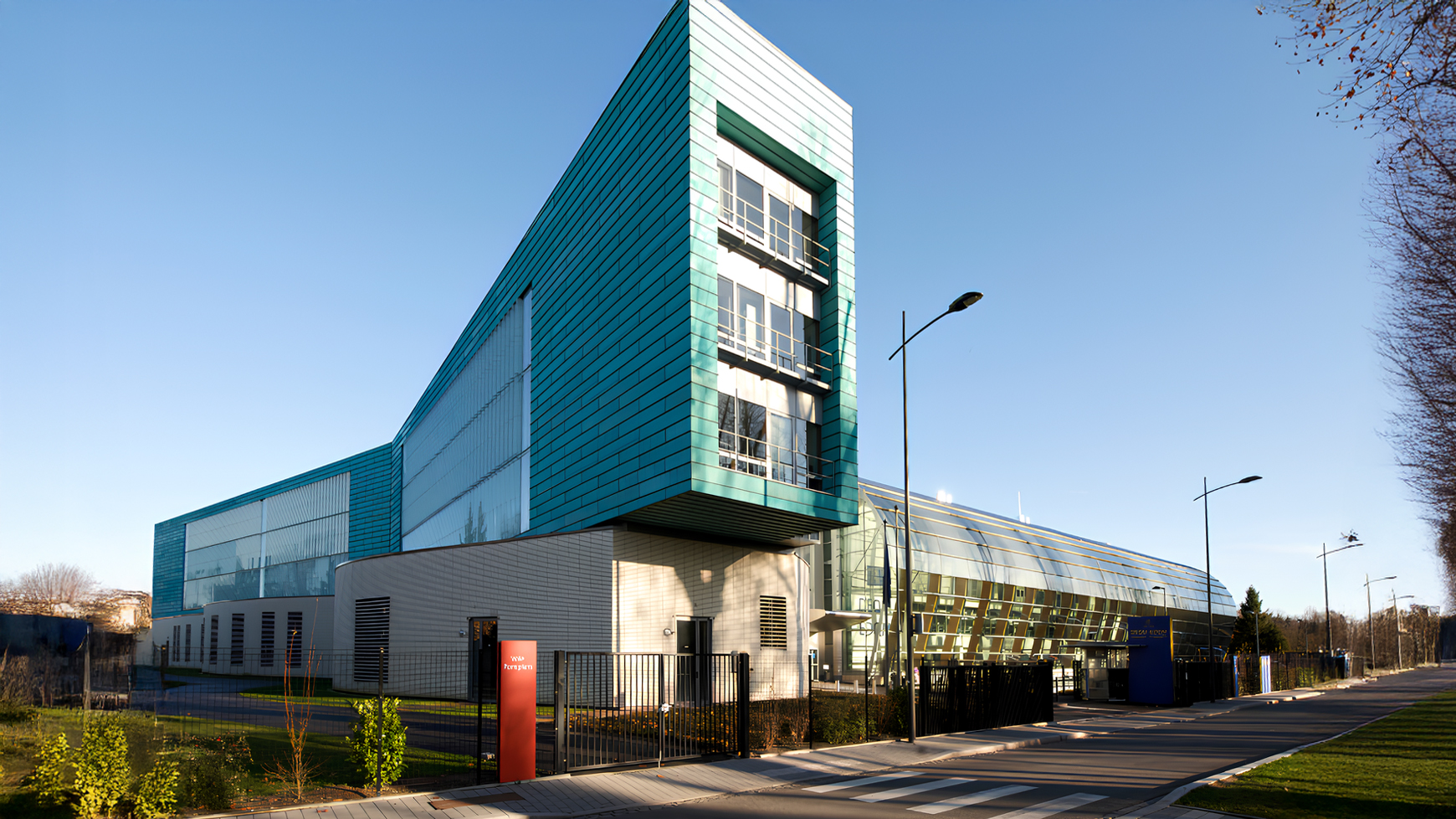 Perspective view of the EDQM building in Strasbourg, featuring a glass façade and aqua-green metal cladding, located along a tree-lined avenue with contemporary street lighting.