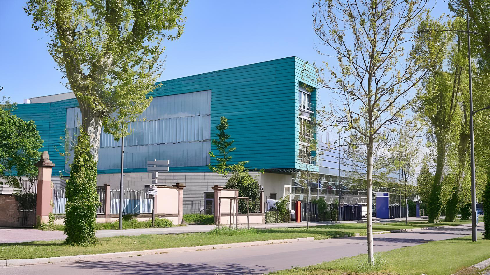 Side view of the EDQM building in Strasbourg, featuring aqua-green panel cladding and large glazed surfaces, surrounded by trees and green spaces along the urban avenue.