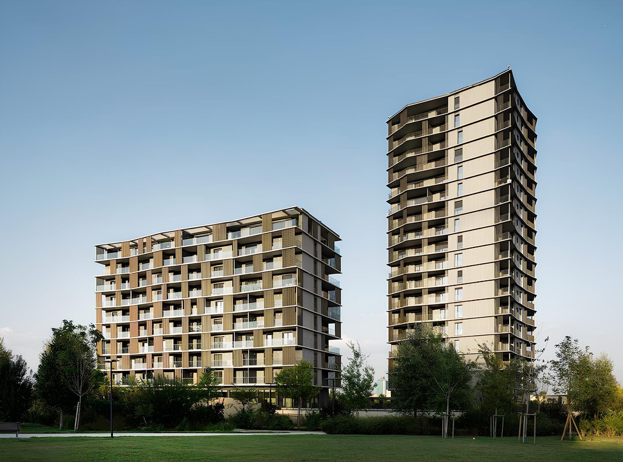 East Uptown, Milano. Due edifici residenziali contemporanei di diversa altezza, caratterizzati da facciate modulari e logge aggettanti, immersi in un contesto verde urbano con alberi e spazi pubblici aperti.