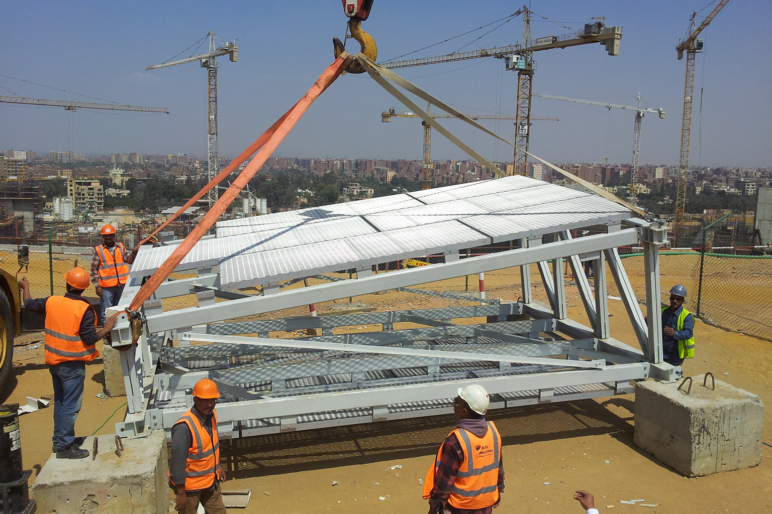 Il Cairo, Egypt. Installation operation of a prefabricated structural module from the GEM project, with on-site workers equipped with safety gear and cranes operating in an urban development area.