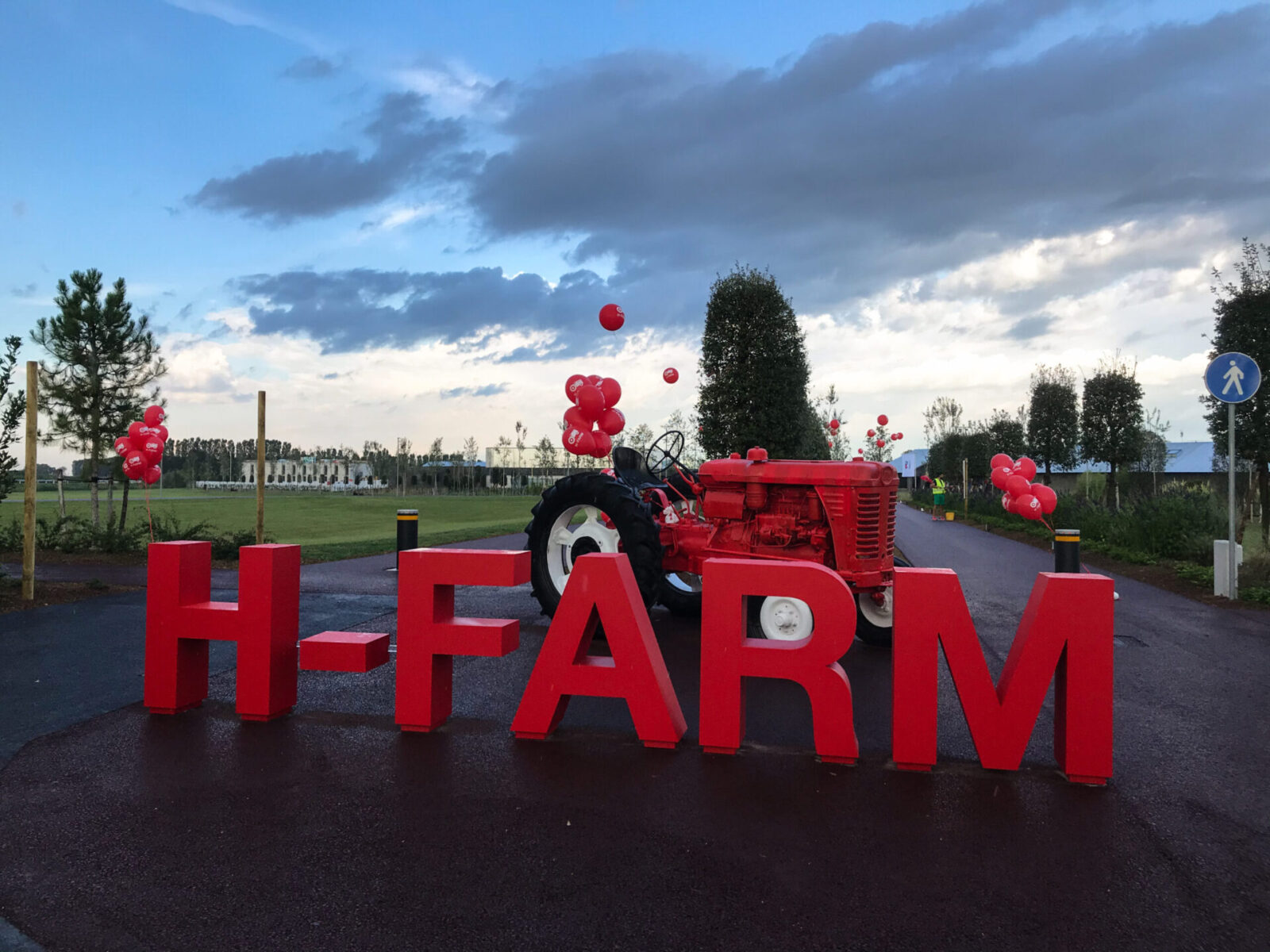 H-Campus, Roncade. Scenic entrance of H-FARM featuring large red 3D letters and a vintage red tractor, surrounded by red balloons. The setup is placed along a tree-lined path in a rural setting, with a cloudy sky in the background, symbolizing a blend of innovation, tradition, and nature.