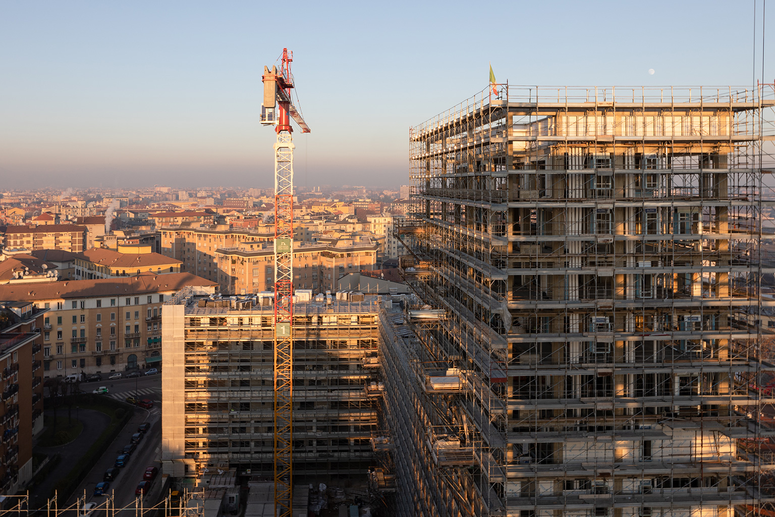 Milano City Village, Milano. Veduta aerea del cantiere MCV alle prime luci del giorno, con edifici in costruzione avvolti da ponteggi, una gru in primo piano e sullo sfondo il tessuto urbano milanese.