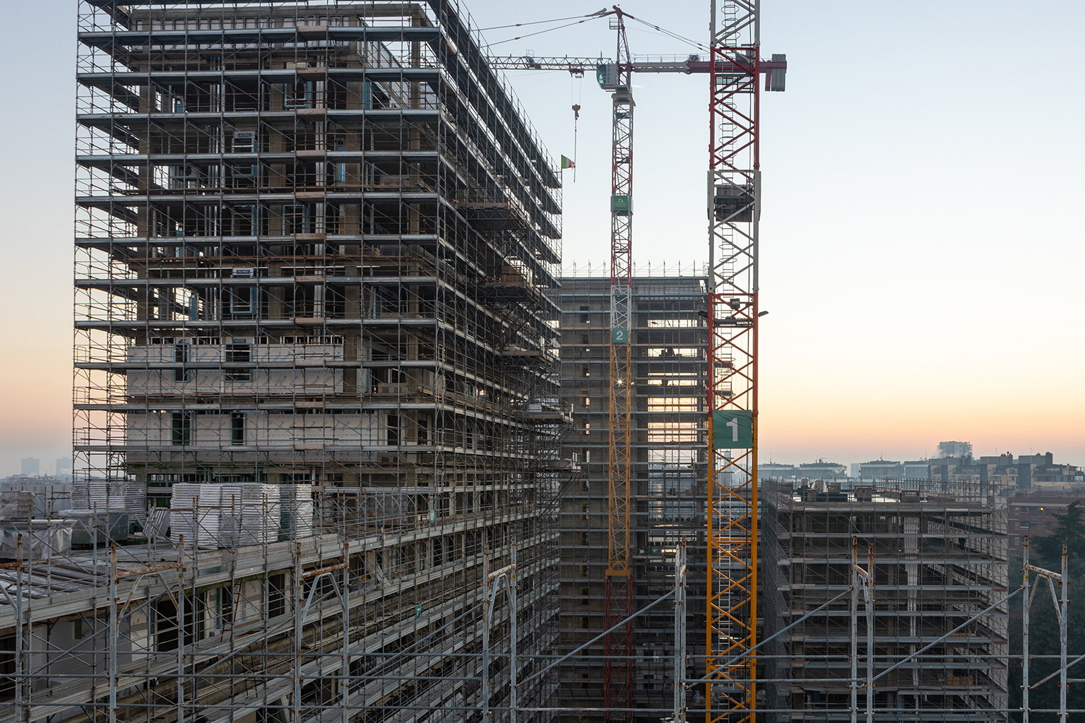 Milano City Village, Milano. Vista panoramica del cantiere MCV al tramonto, con edifici in elevazione avvolti da impalcature, gru da cantiere in funzione e skyline urbano sullo sfondo.