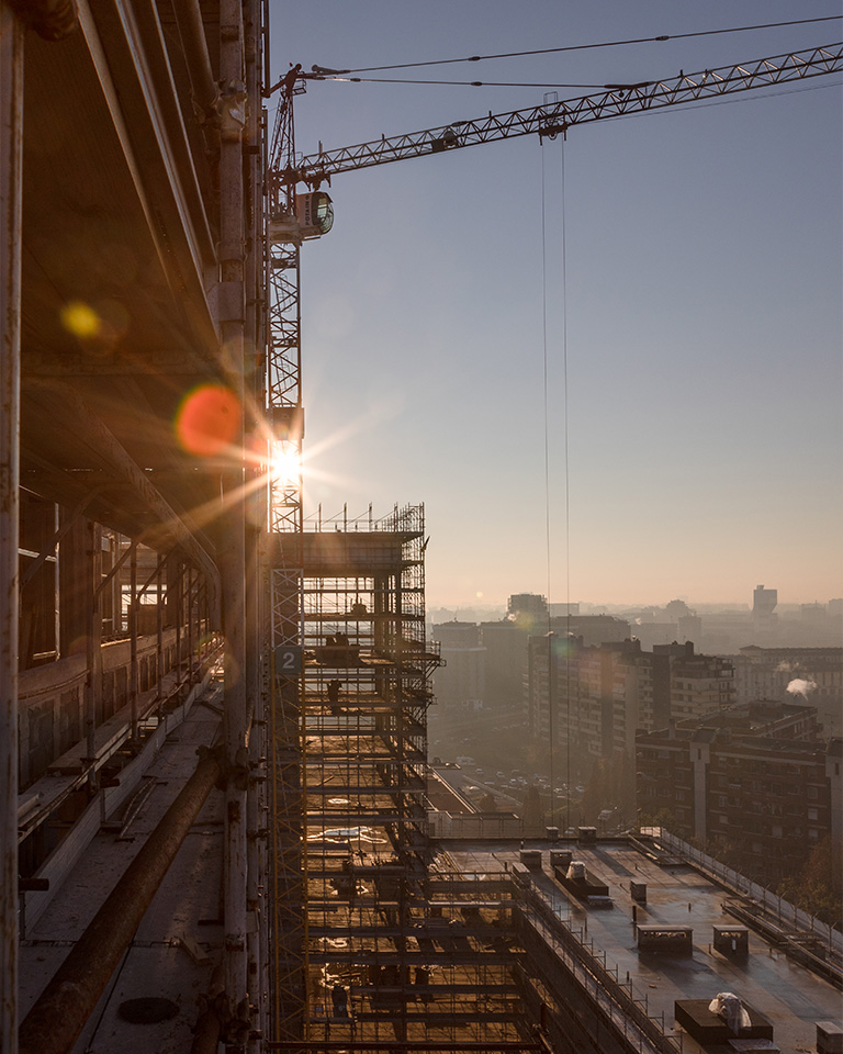 Milano City Village, Milano. Scorcio del cantiere MCV alle prime luci del mattino, con impalcature illuminate dal sole, gru in primo piano e vista urbana immersa nella foschia.