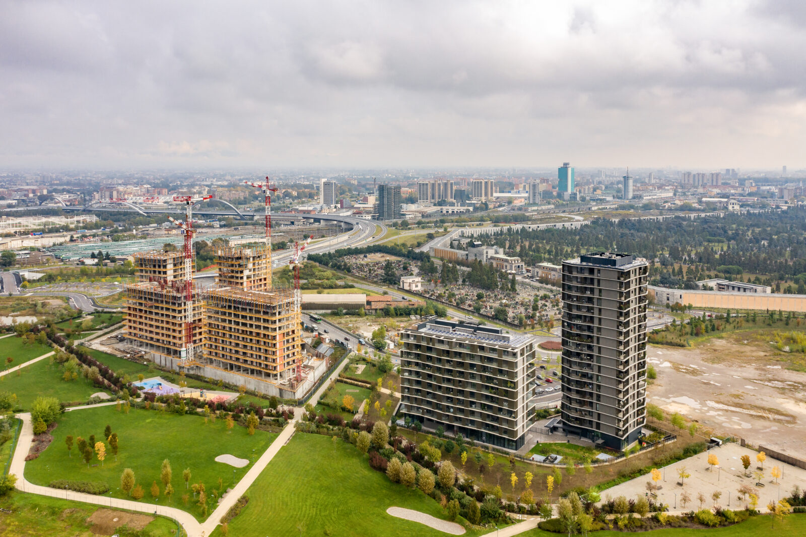 Aerial view of the Cascina Merlata district in Milan, featuring residential buildings under construction alongside completed structures, all surrounded by a large urban park. The city skyline appears in the background, intersected by major roads and railway lines.