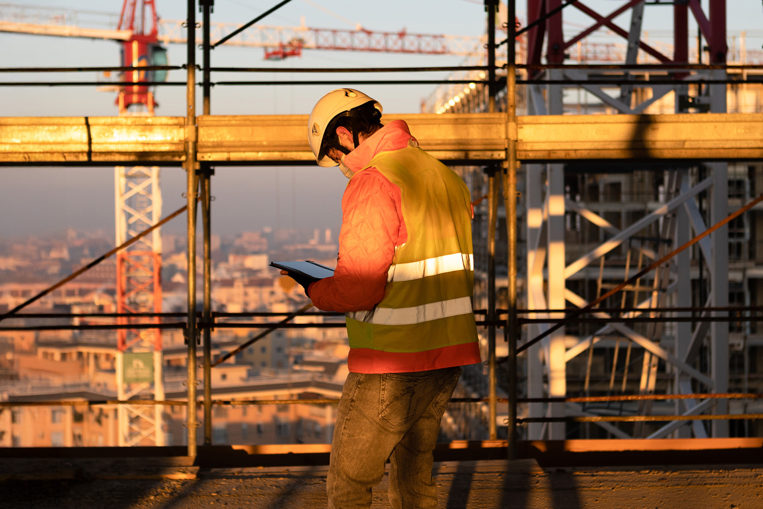 Milano City Viillage, Milan. Construction technician wearing a helmet and high-visibility vest checks a tablet at a building site during sunset, with scaffolding and cranes in the background.