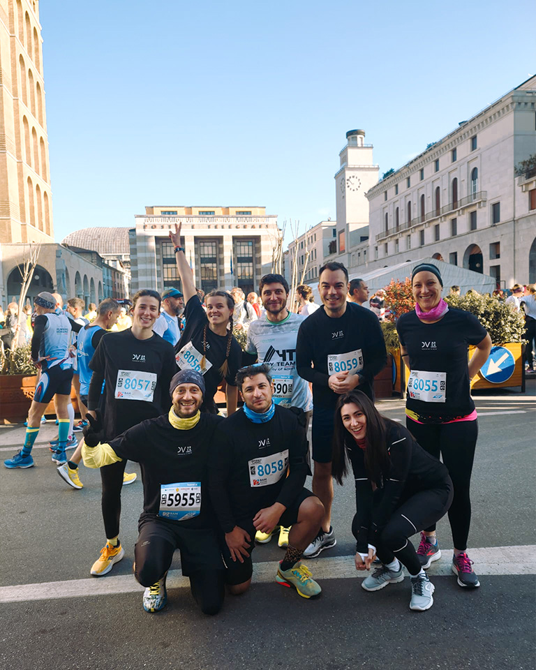 Brescia Art Marathon 2023. Un gruppo di dipendenti DVArea posa per una foto prima di una corsa podistica cittadina, indossando abbigliamento sportivo e pettorine numerate. Alle loro spalle, altri partecipanti e palazzi storici in una piazza assolata.