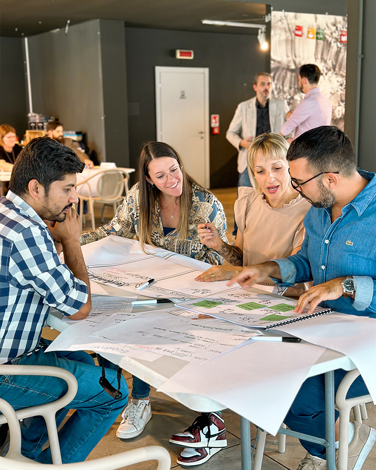 Four people actively collaborate during a workshop, discussing diagrams and notes spread out on a large table; in the background, other participants engage in an open, modern space decorated with references to the Sustainable Development Goals.