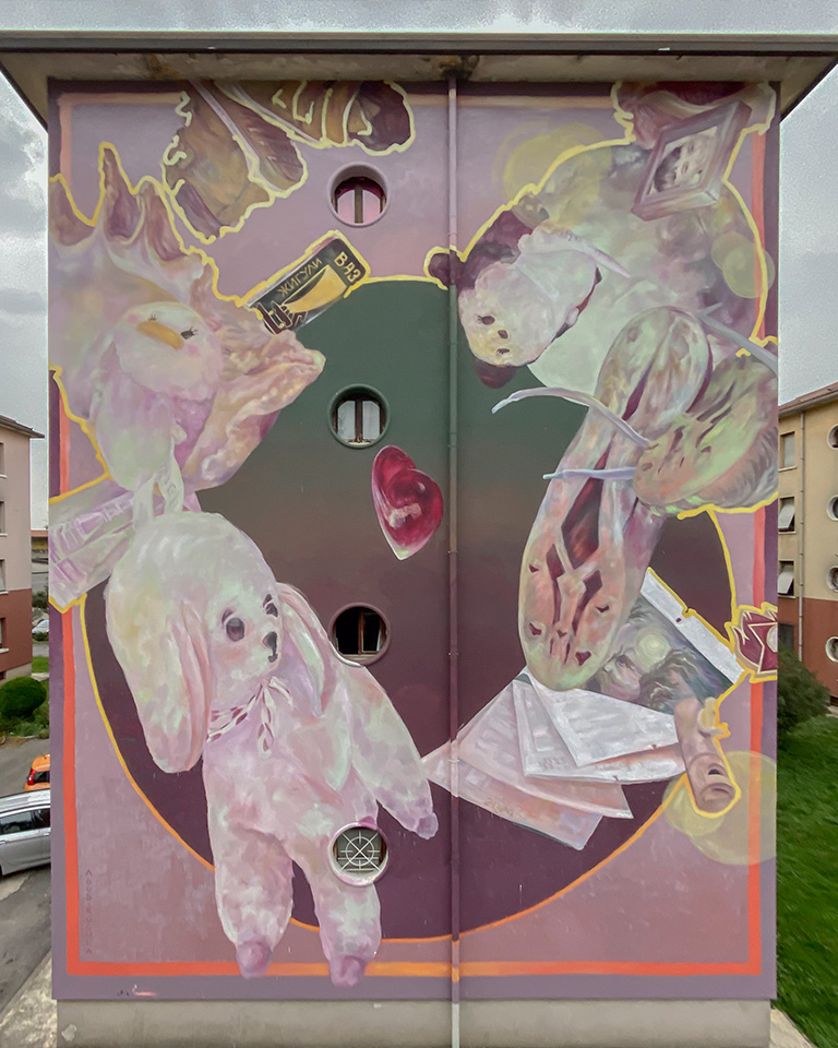 Grande murale dipinto sulla facciata di un edificio residenziale, raffigurante una scena onirica e surreale con pupazzi, oggetti quotidiani, libri e un cuore rosso fluttuante, il tutto immerso in tonalità pastello tra rosa, viola e verde scuro, con dettagli sfumati e contorni dorati.