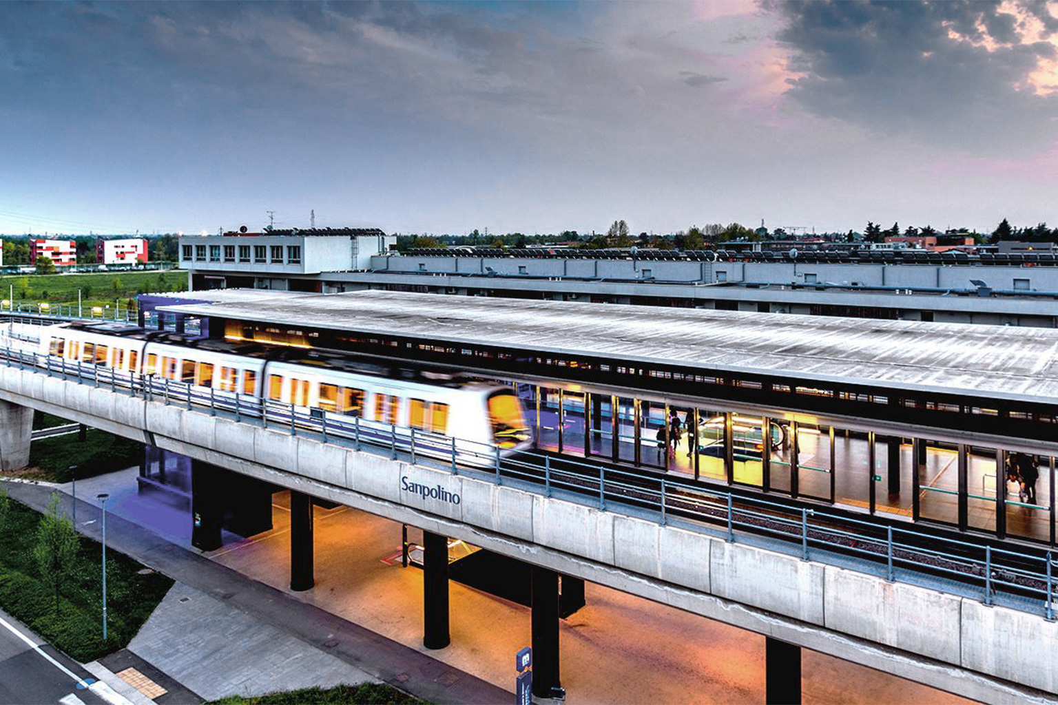 Brescia Infrastrutture, Brescia. Evening view of the elevated “Sanpolino” station on the automated metro line in Brescia. A moving train passes through the lit platform, with warm reflections and interior lights visible through the glass walls. In the background, industrial and residential buildings are silhouetted under a cloudy sunset sky.