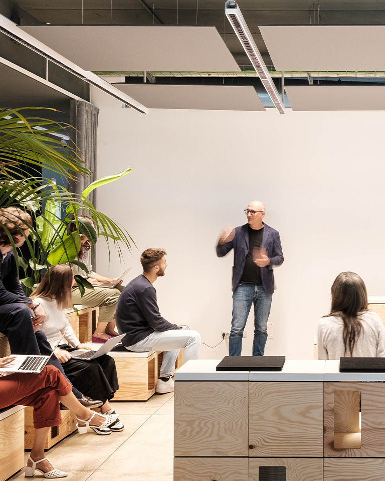 Armando Casella stands while speaking to a group of colleagues seated on modular wooden steps, some with laptops, in an informal room featuring light furnishings, plants, and technical lighting.