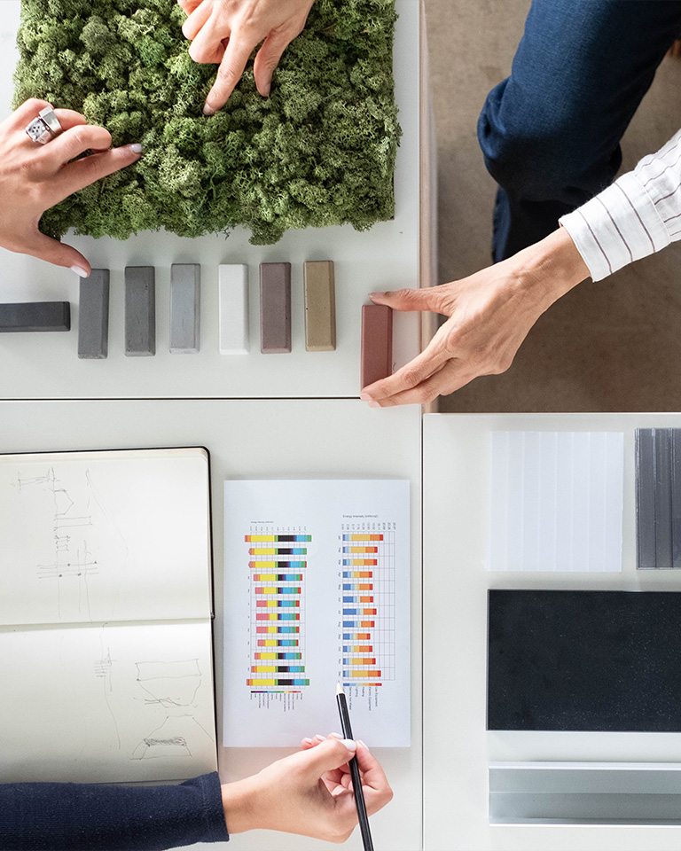 Top-down view of a worktable with hands interacting with material samples, including colored swatches, translucent panels, multicolored bar charts, design sketches, and a green moss panel, in an architectural or interior design setting.