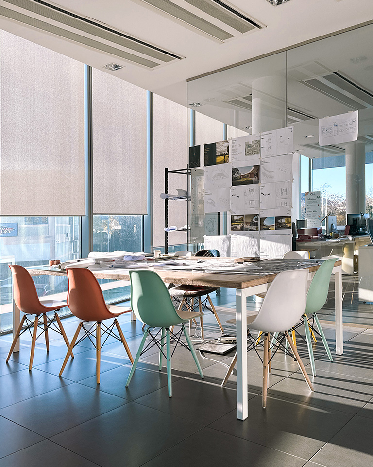 Spazio di lavoro creativo con tavolo in legno, sedie colorate e parete vetrata con disegni tecnici esposti.