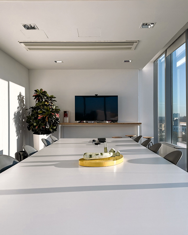 Sala riunioni moderna e luminosa con ampie vetrate, un lungo tavolo bianco centrale, sedie grigie, uno schermo TV montato a parete e un modellino architettonico posizionato al centro del tavolo; pianta decorativa nell’angolo sinistro.