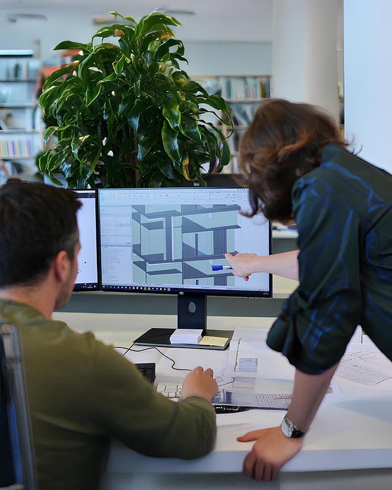 Professionals working on a 3D architectural model using BIM software, displayed on dual monitors in a technical office setting.