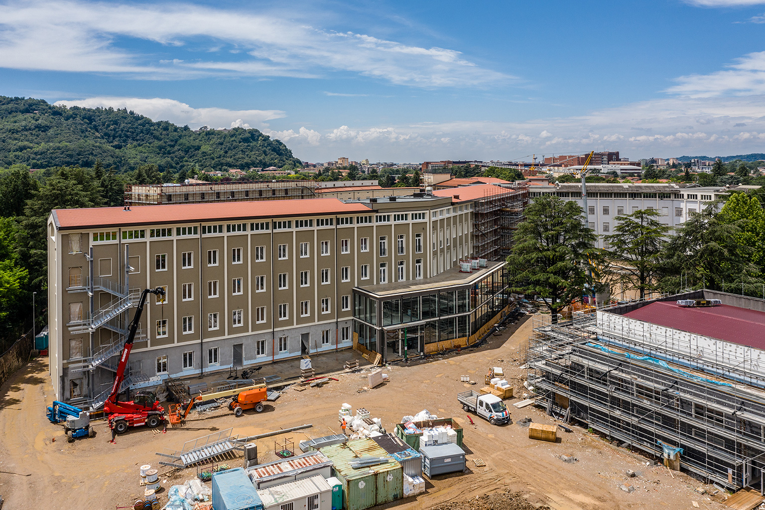 Campus Cattolica, Brescia. Aerial view of the Cattolica complex construction site in an advanced stage of renovation, with restored buildings, scaffolding, operational machinery, and a hilly backdrop.