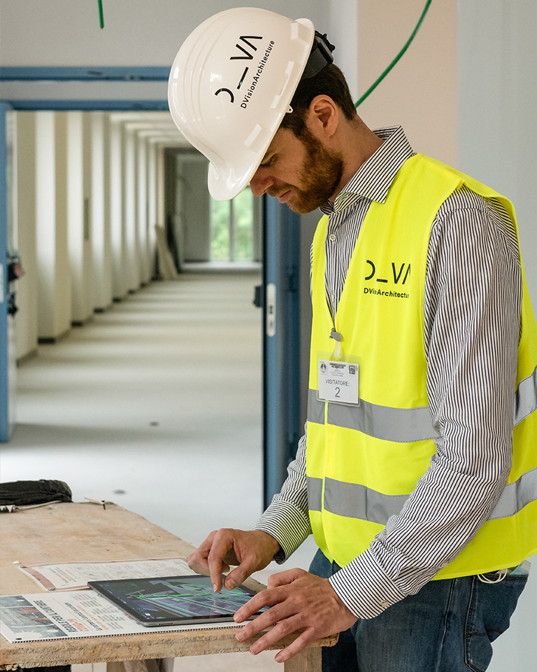 Campus Cattolica, Brescia. DVA technician during an on-site inspection, wearing a hard hat and high-visibility vest, consulting a digital model on a tablet inside a building under construction.