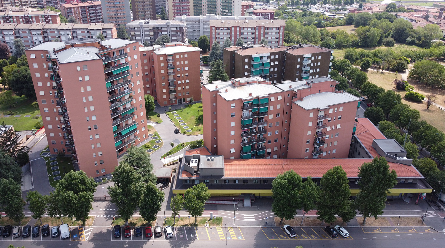 Eleonora Duse, Sesto San Giovanni, Milan. Aerial view of a residential complex composed of multiple mid-rise apartment buildings with red façades and balconies, surrounded by trees, green courtyards, and urban infrastructure in a densely built neighborhood.