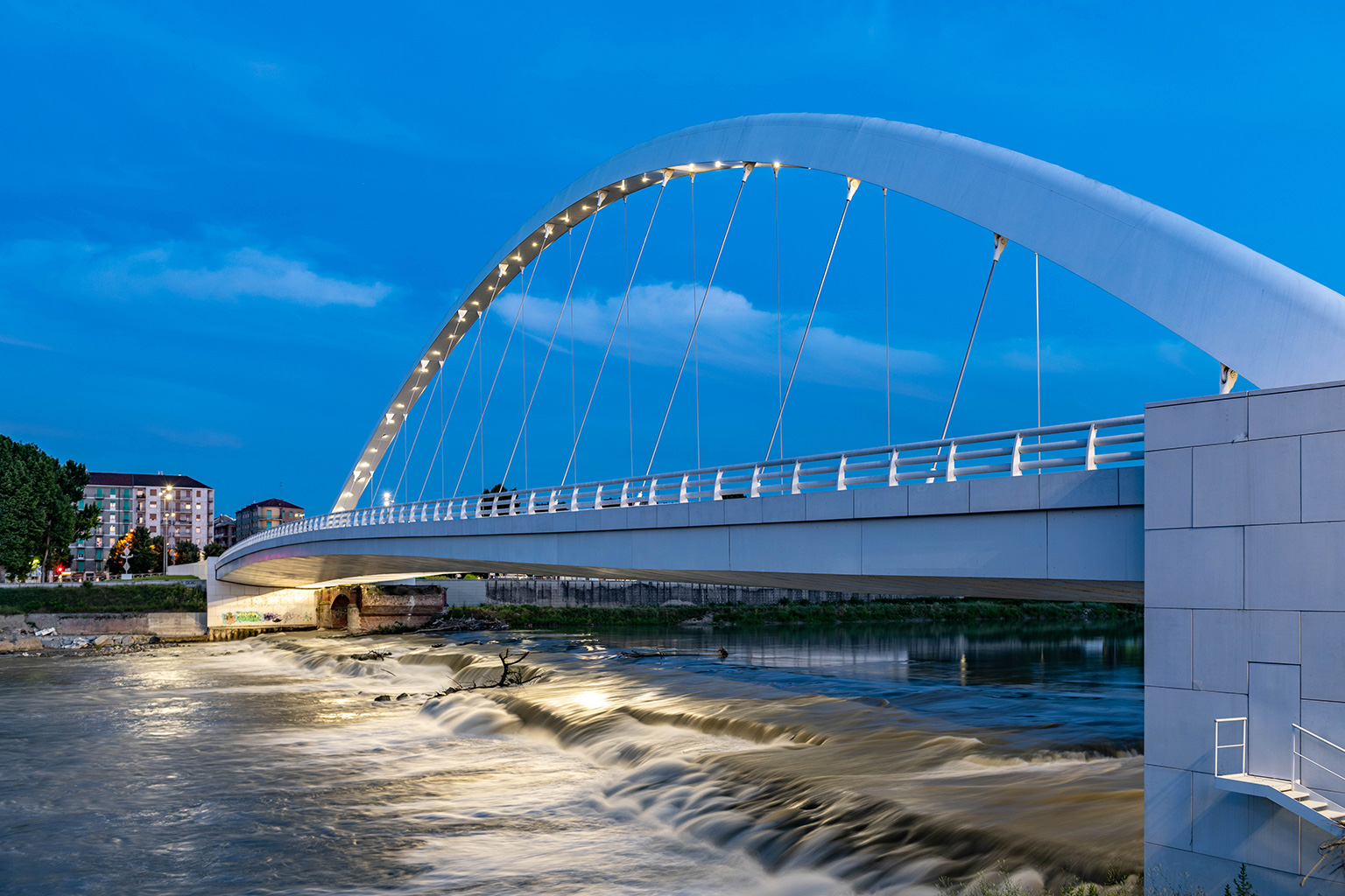 Cittadella Bridge, Alessandria. Evening view of the Cittadella Bridge in Alessandria, featuring a backlit white arch and metallic cables reflecting on the river below, framed by a twilight blue sky and an urban backdrop.